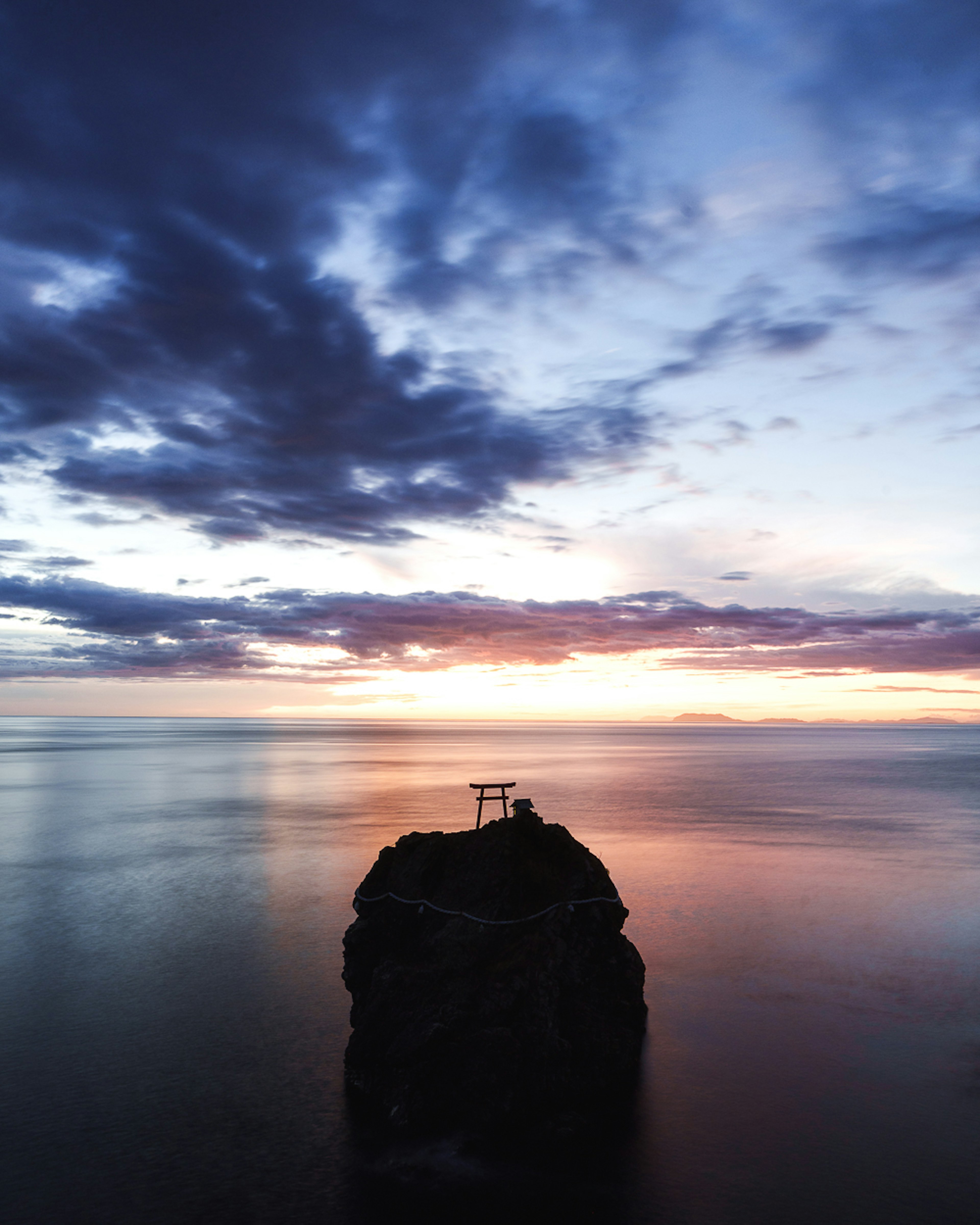 Un coucher de soleil serein sur des eaux calmes avec un rocher et une porte torii