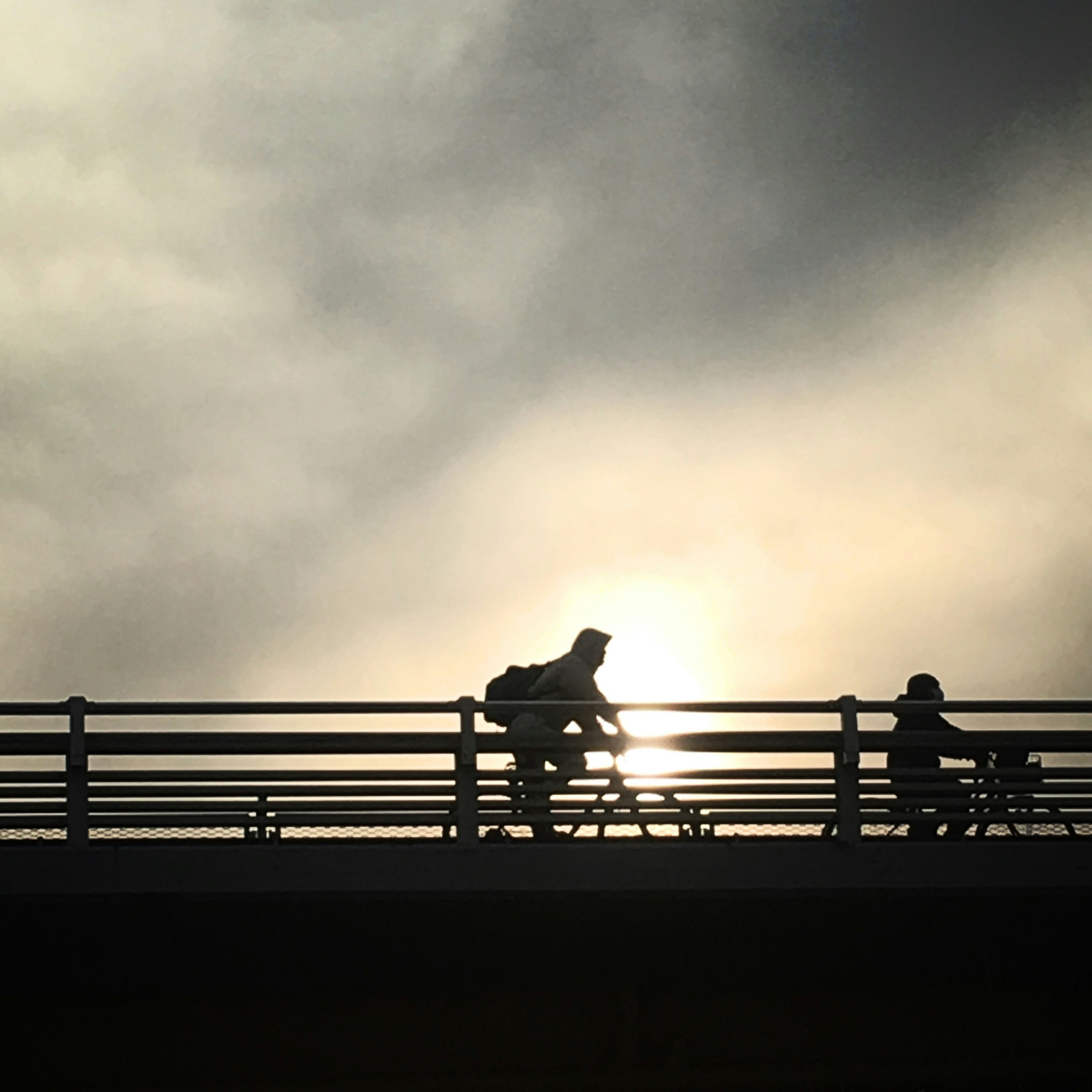 Silhouette von zwei Radfahrern auf einer Brücke vor einem bewölkten Himmel