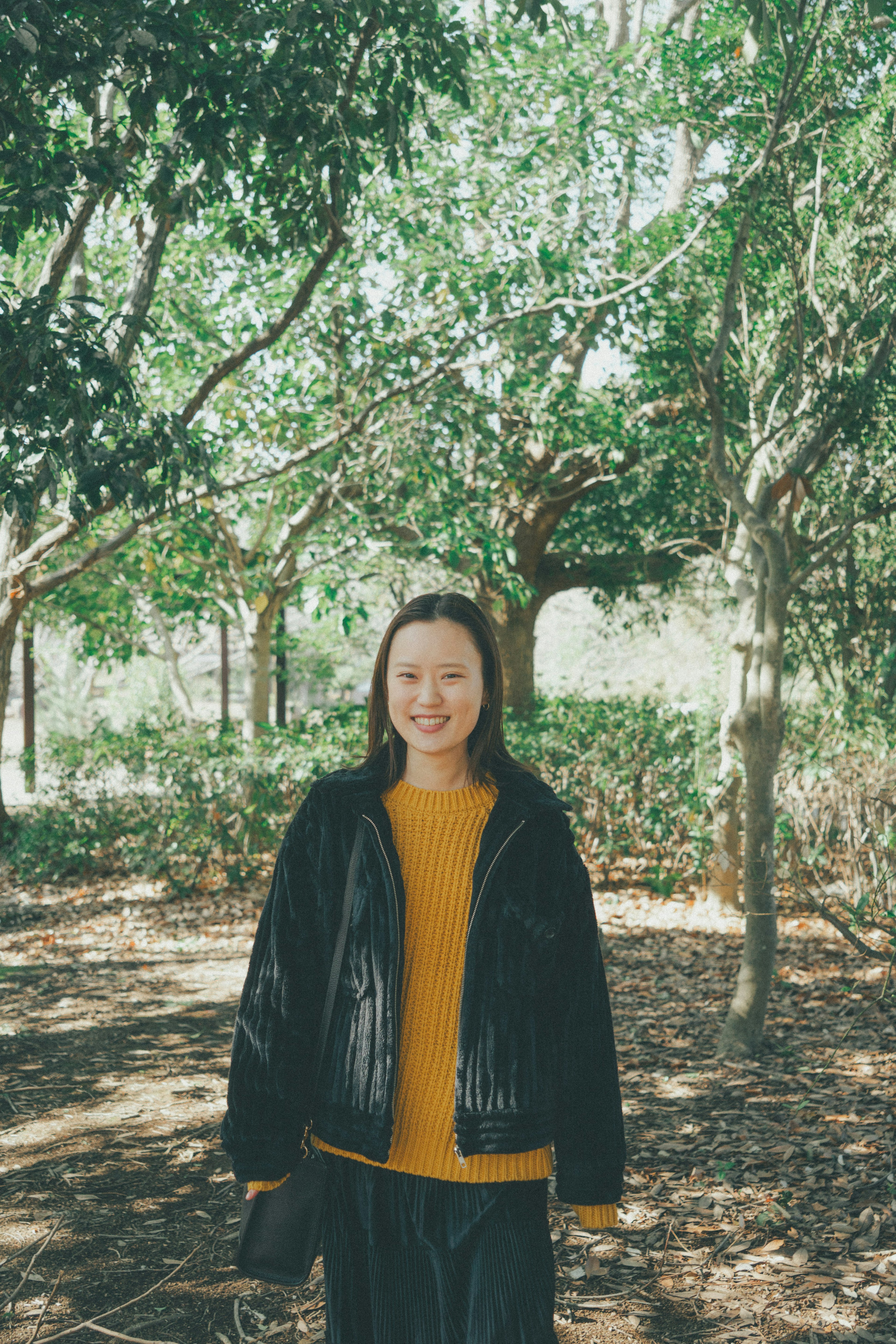 A woman standing among trees smiling wearing a black jacket and an orange sweater