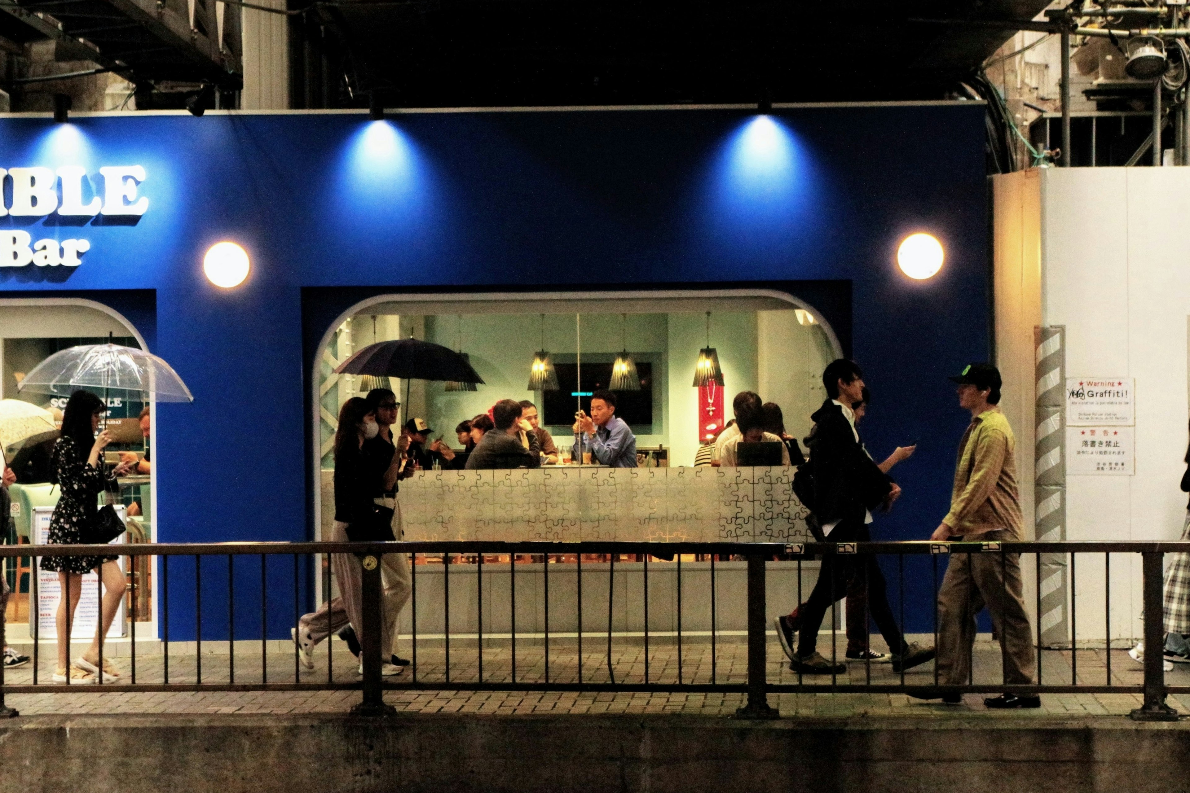 Außenansicht einer blauen Bar mit Menschen, die unter Regenschirmen gehen