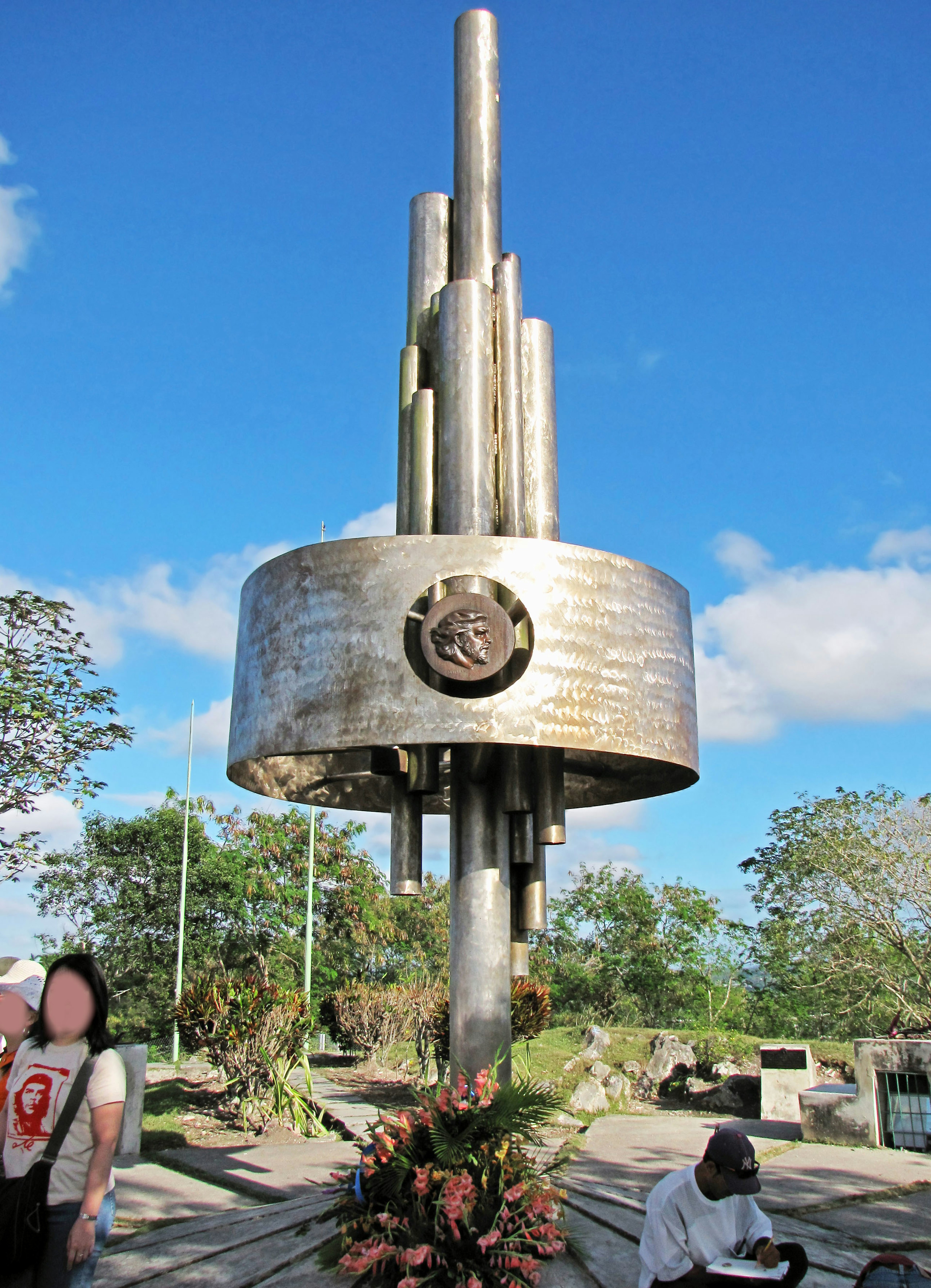 Metal sculpture monument with floral offerings under a blue sky