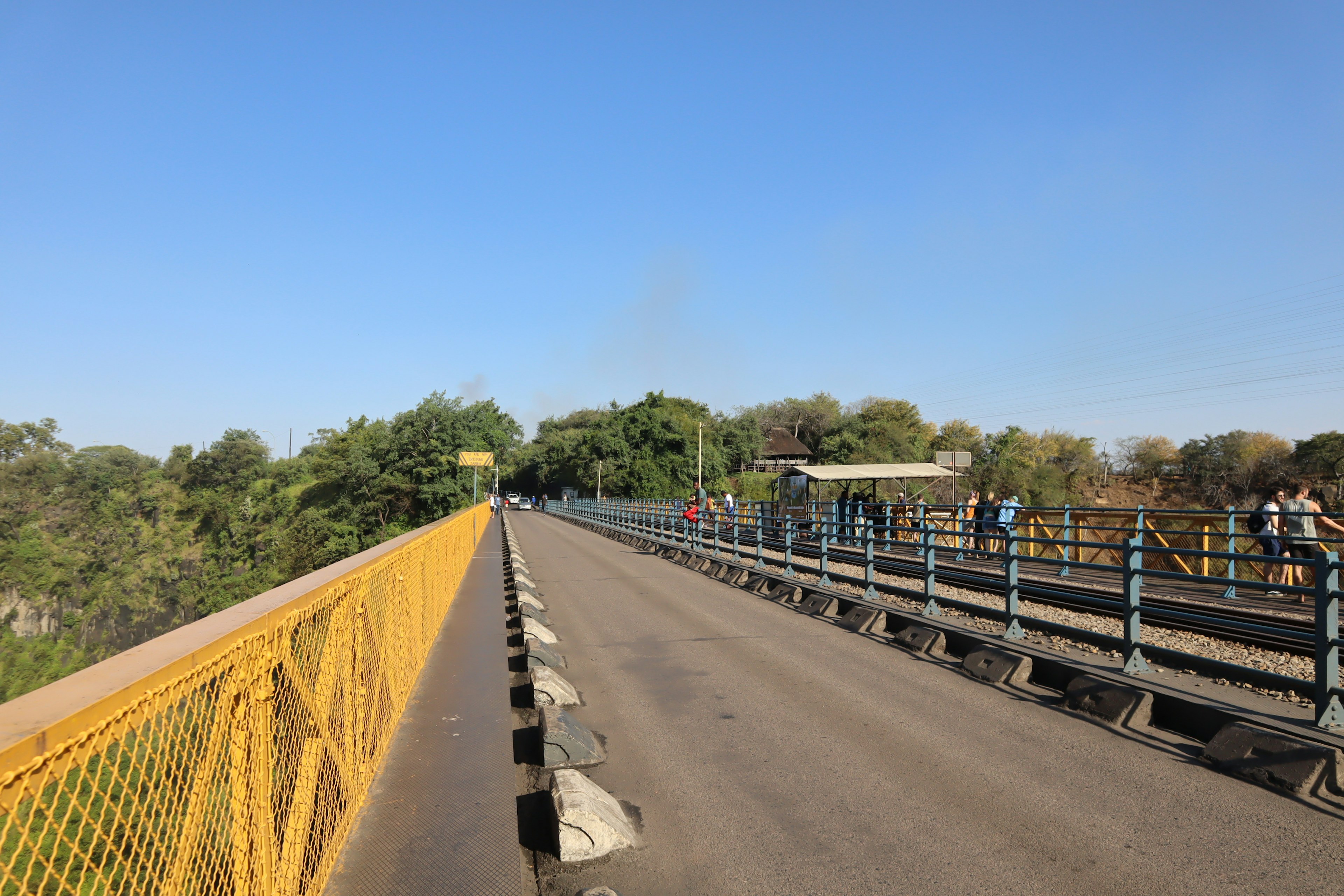 Pemandangan jembatan dengan pagar kuning dikelilingi pohon hijau dan langit biru