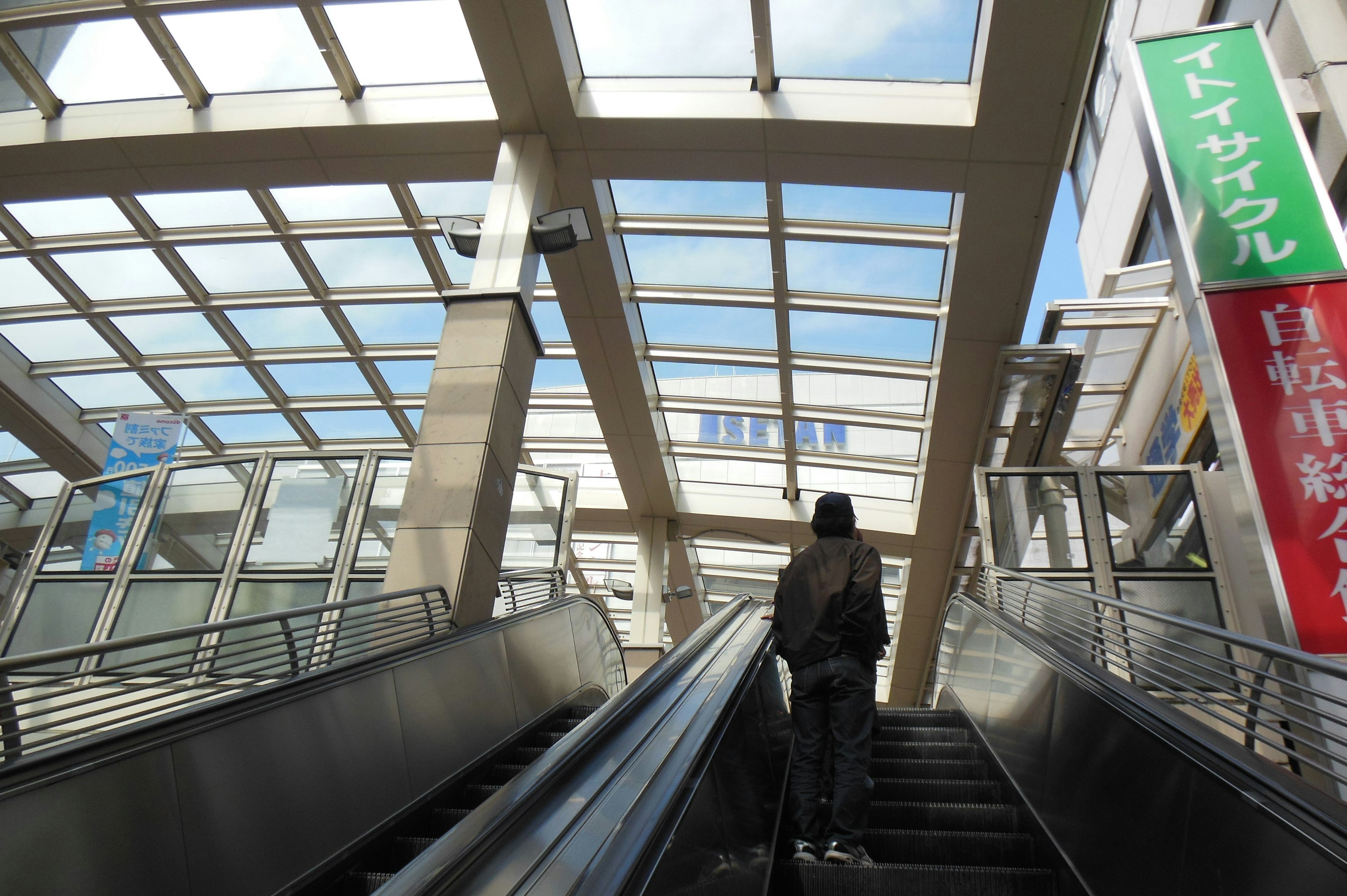 屋外のエスカレーターを上る人々と青空が見える駅の風景