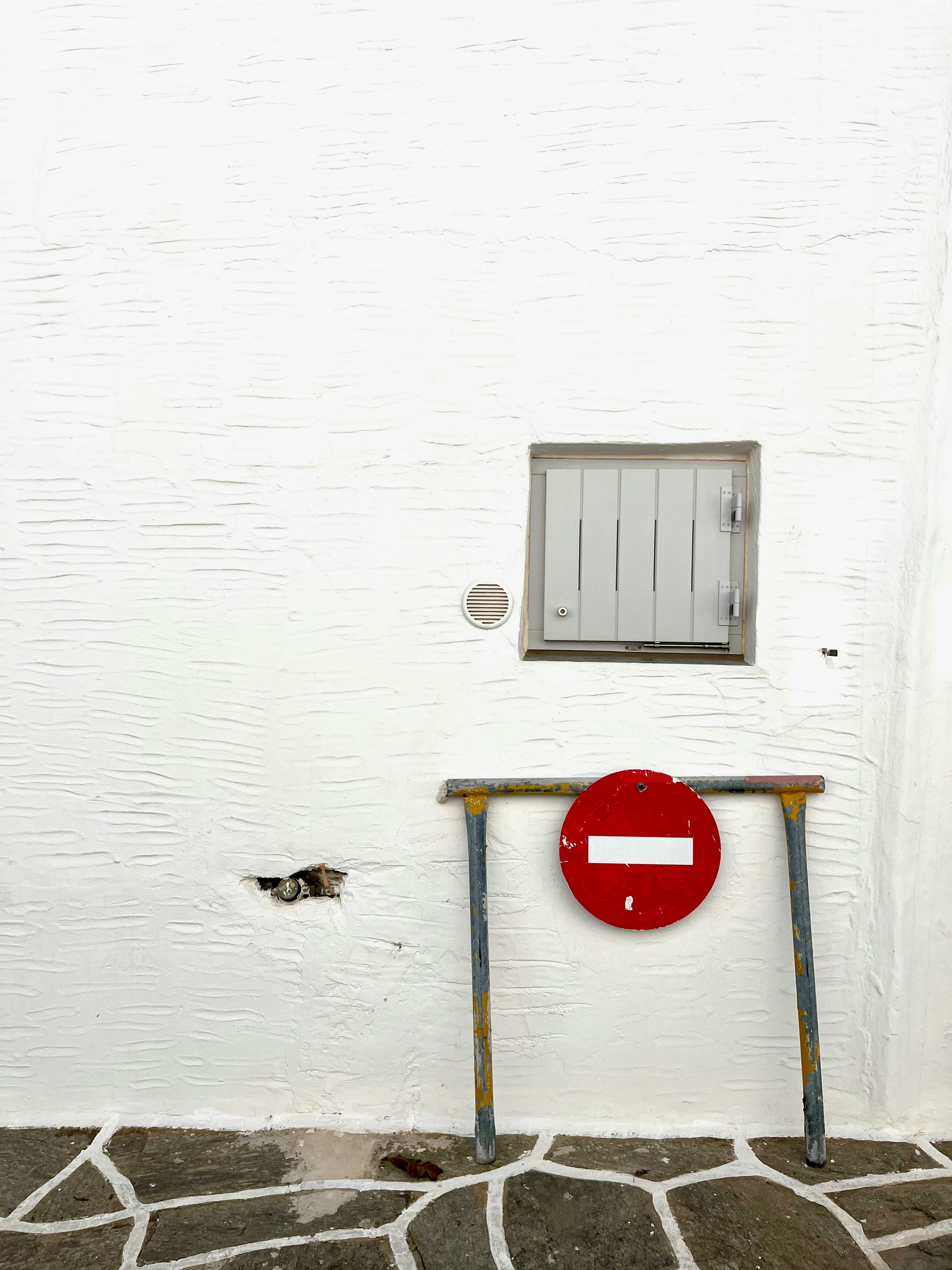 A simple composition featuring a small window and a red no entry sign on a white wall