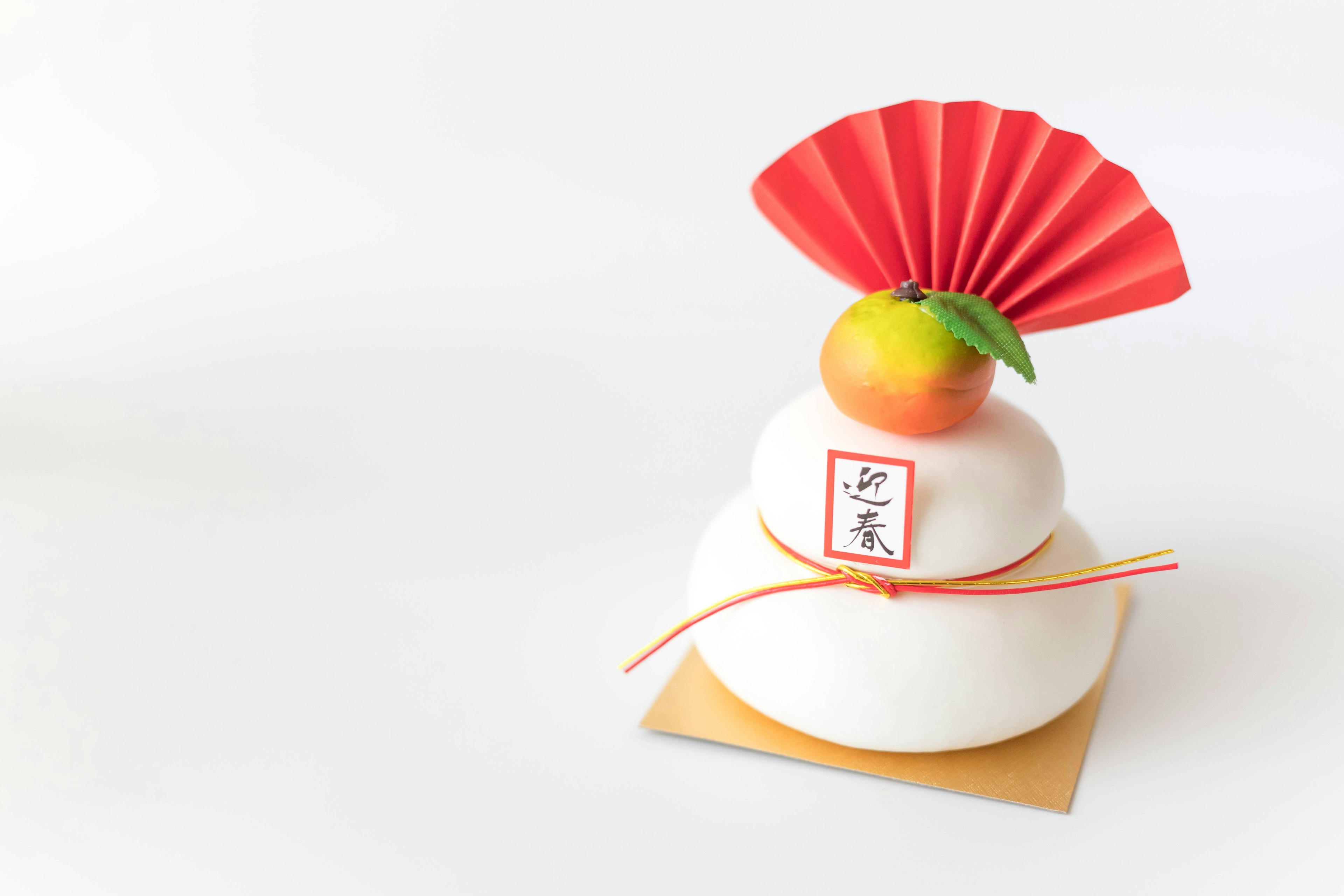 A two-tiered rice cake decoration with a red fan and a mandarin orange on top against a white background