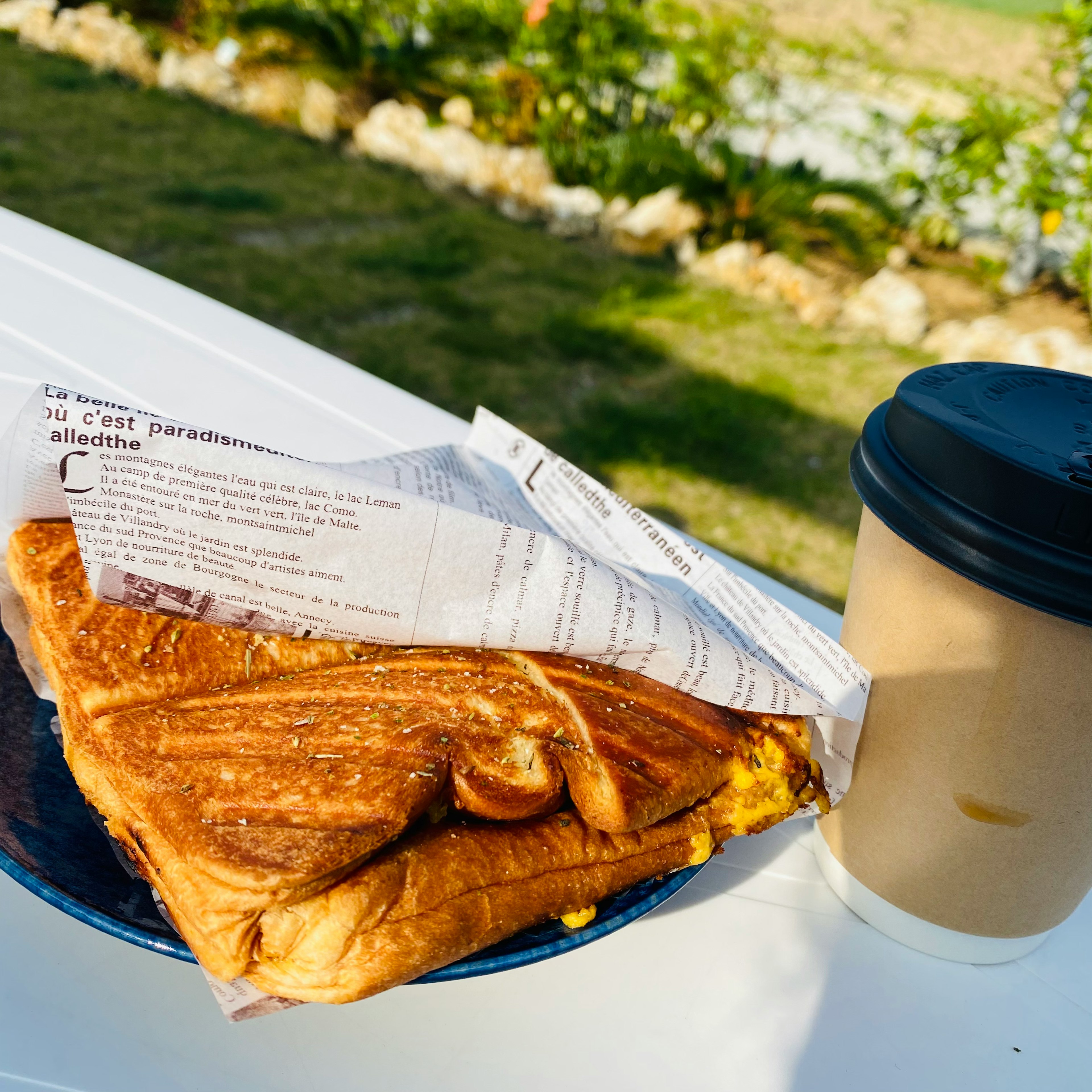 Ein Sandwich auf einem blauen Teller begleitet von einer Kaffeetasse mit schwarzem Deckel vor einem grünen Hintergrund