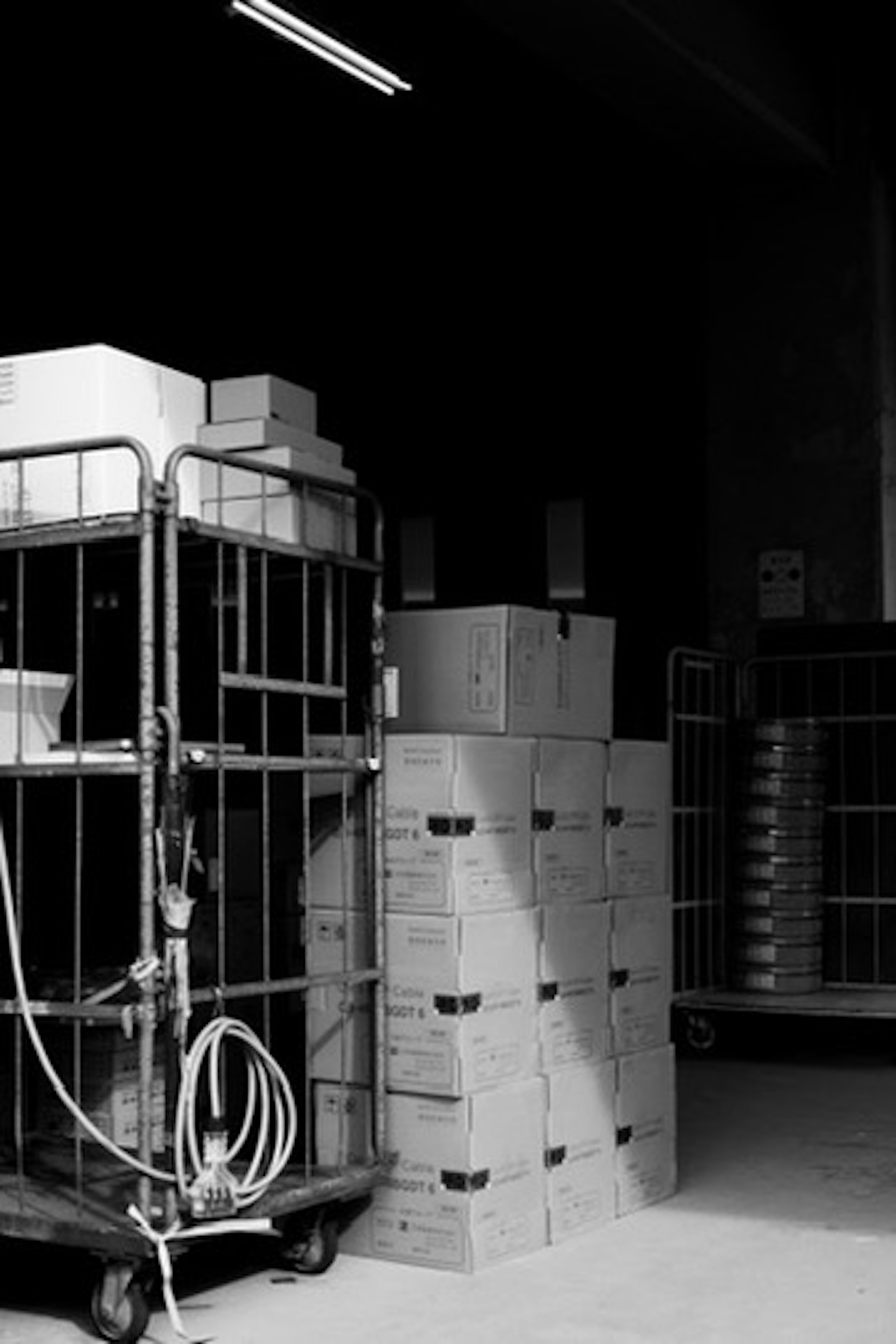 Black and white interior of a warehouse with stacked cardboard boxes and a transport cart