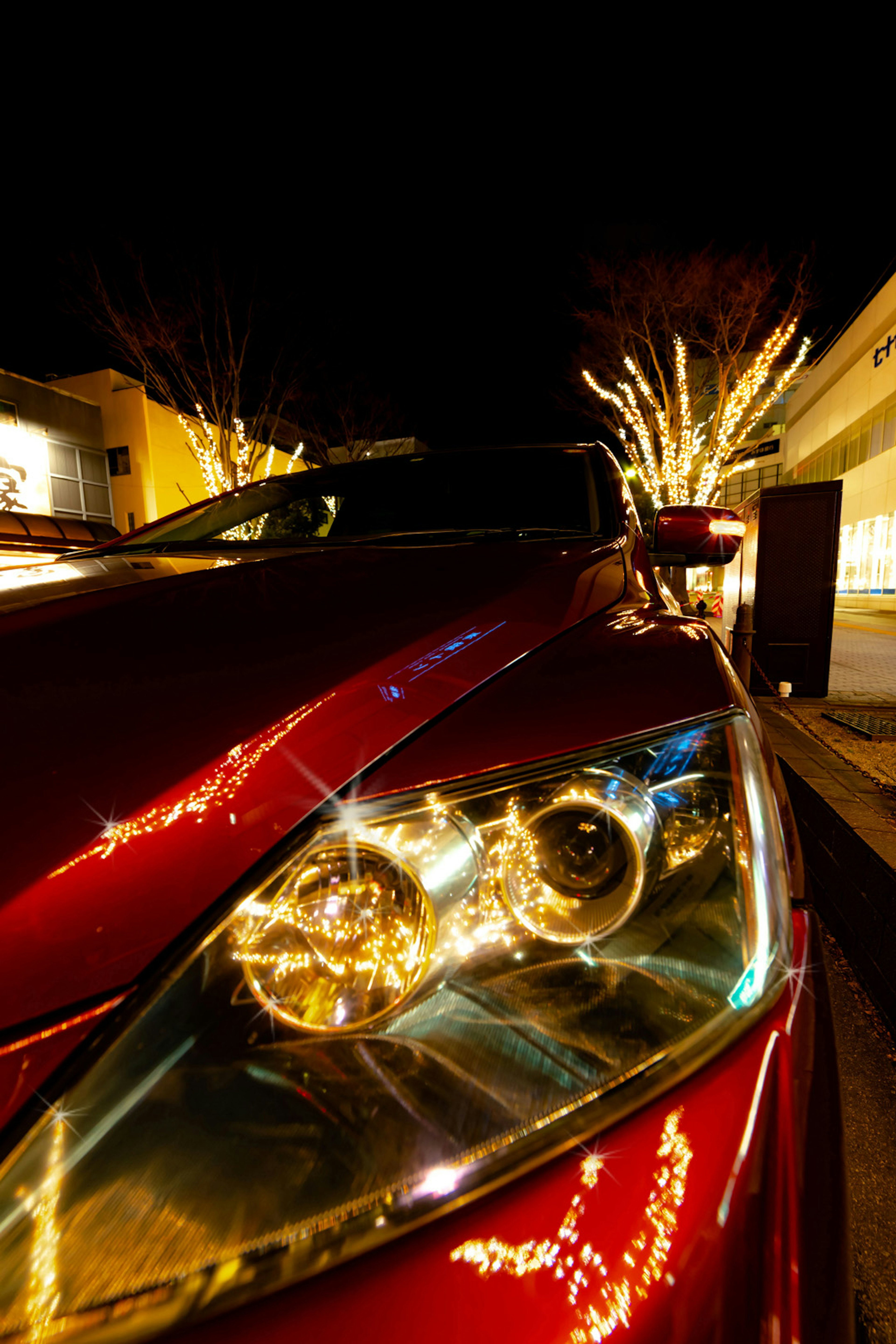 Primo piano dei fari di un'auto rossa illuminata di notte con alberi decorati sullo sfondo