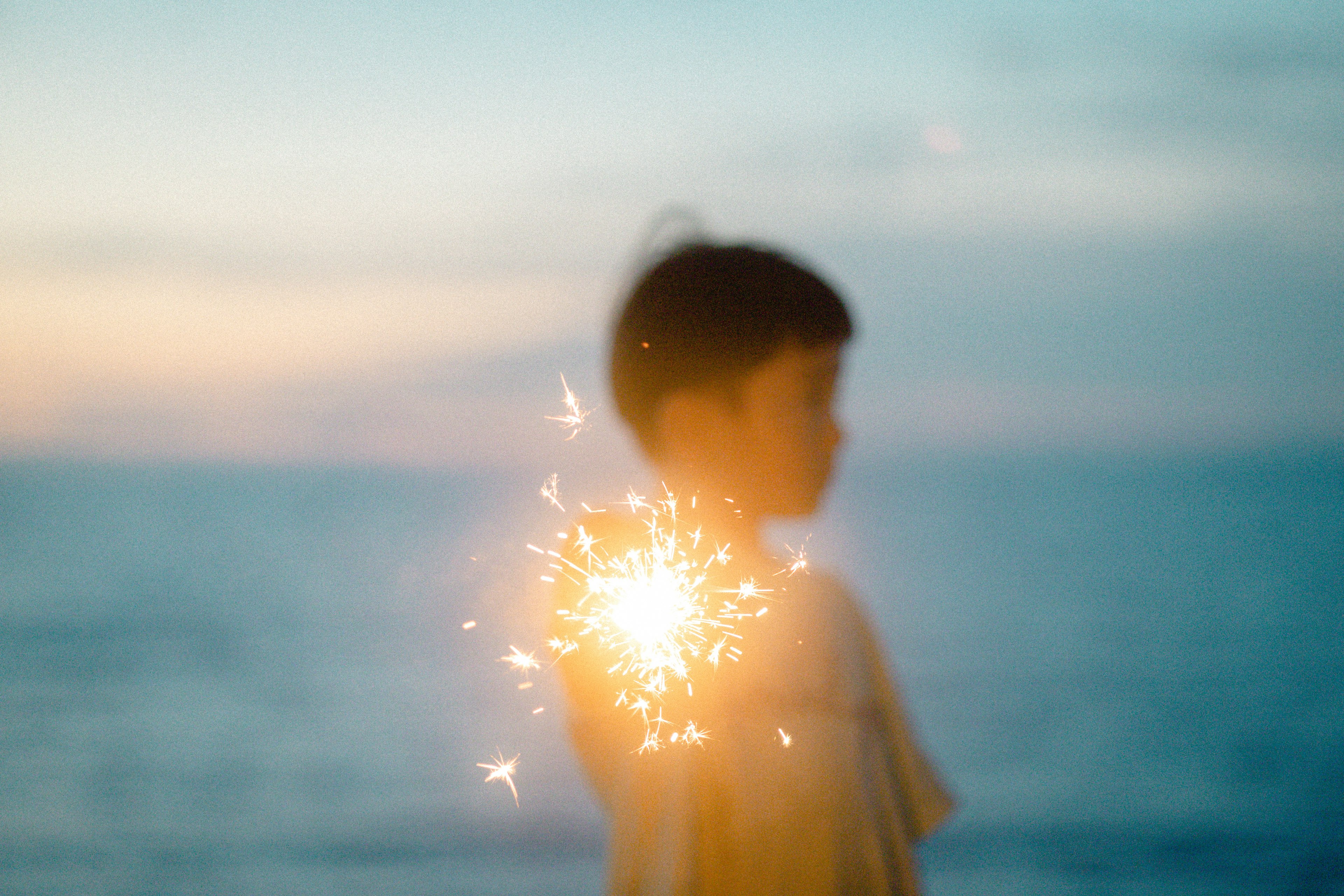 Silhouette seorang anak memegang kembang api di tepi laut saat matahari terbenam