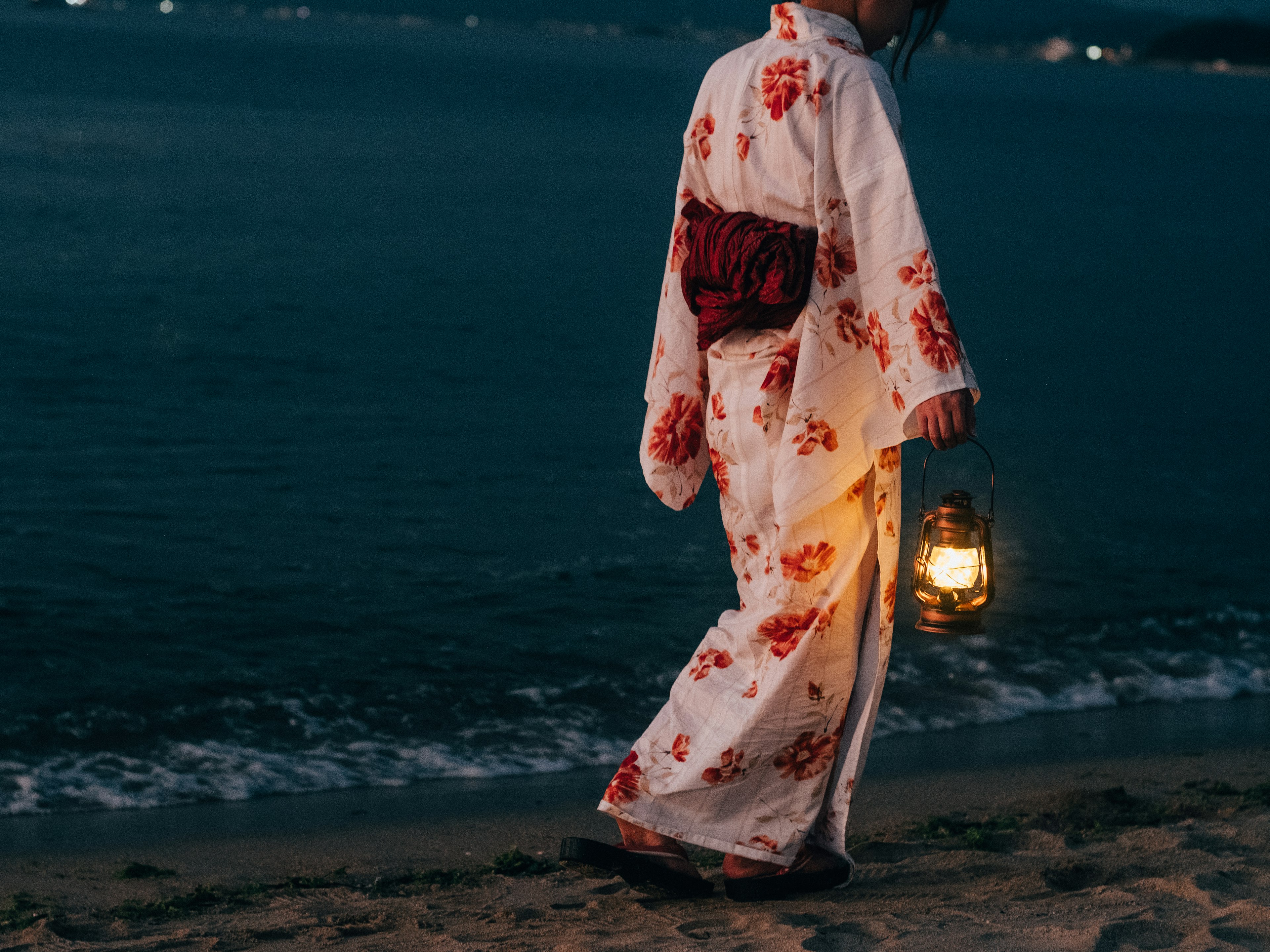 Mujer con kimono floral caminando por la playa sosteniendo una linterna