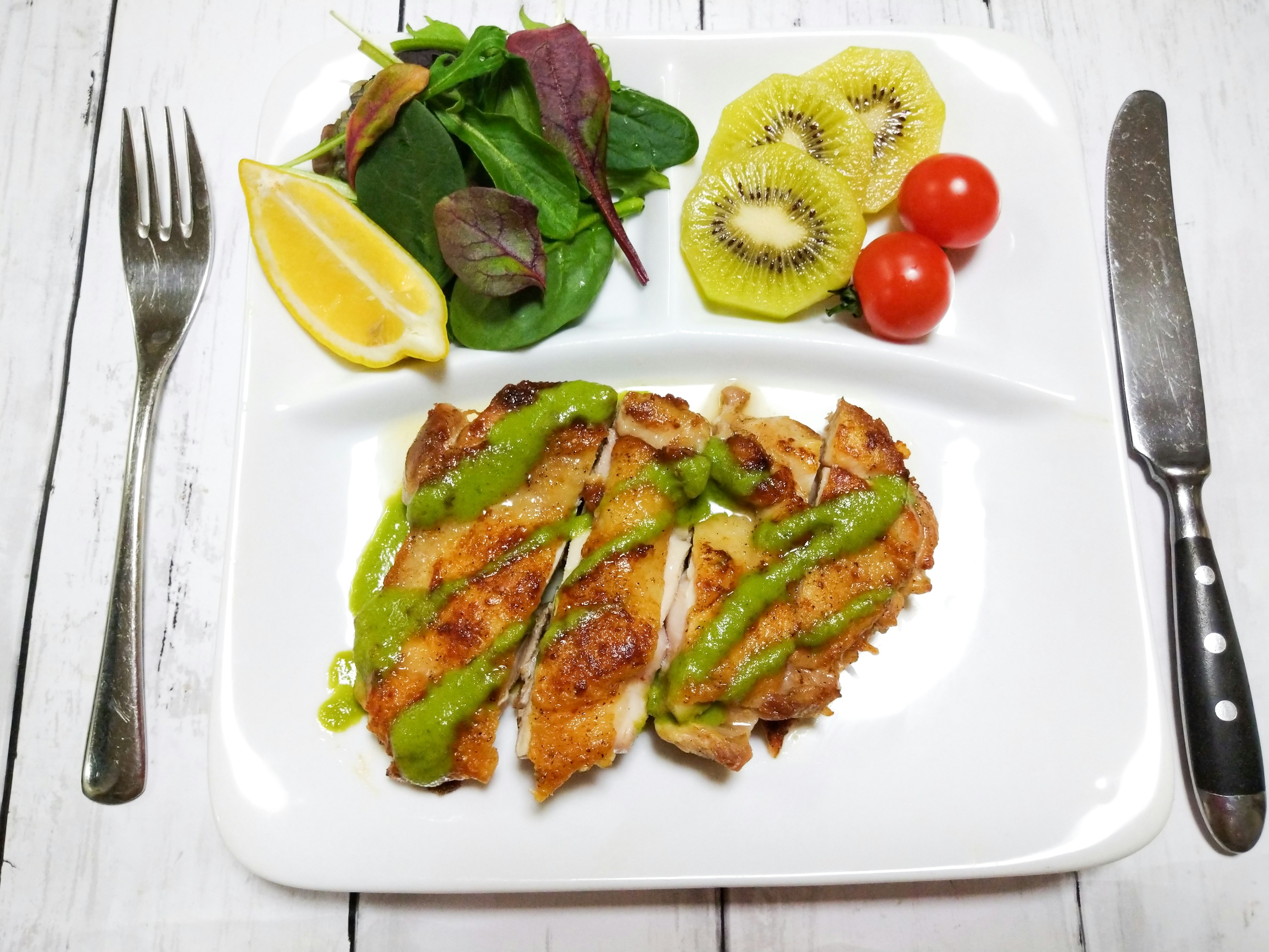 A plate featuring sliced grilled chicken with green sauce alongside salad and kiwi slices