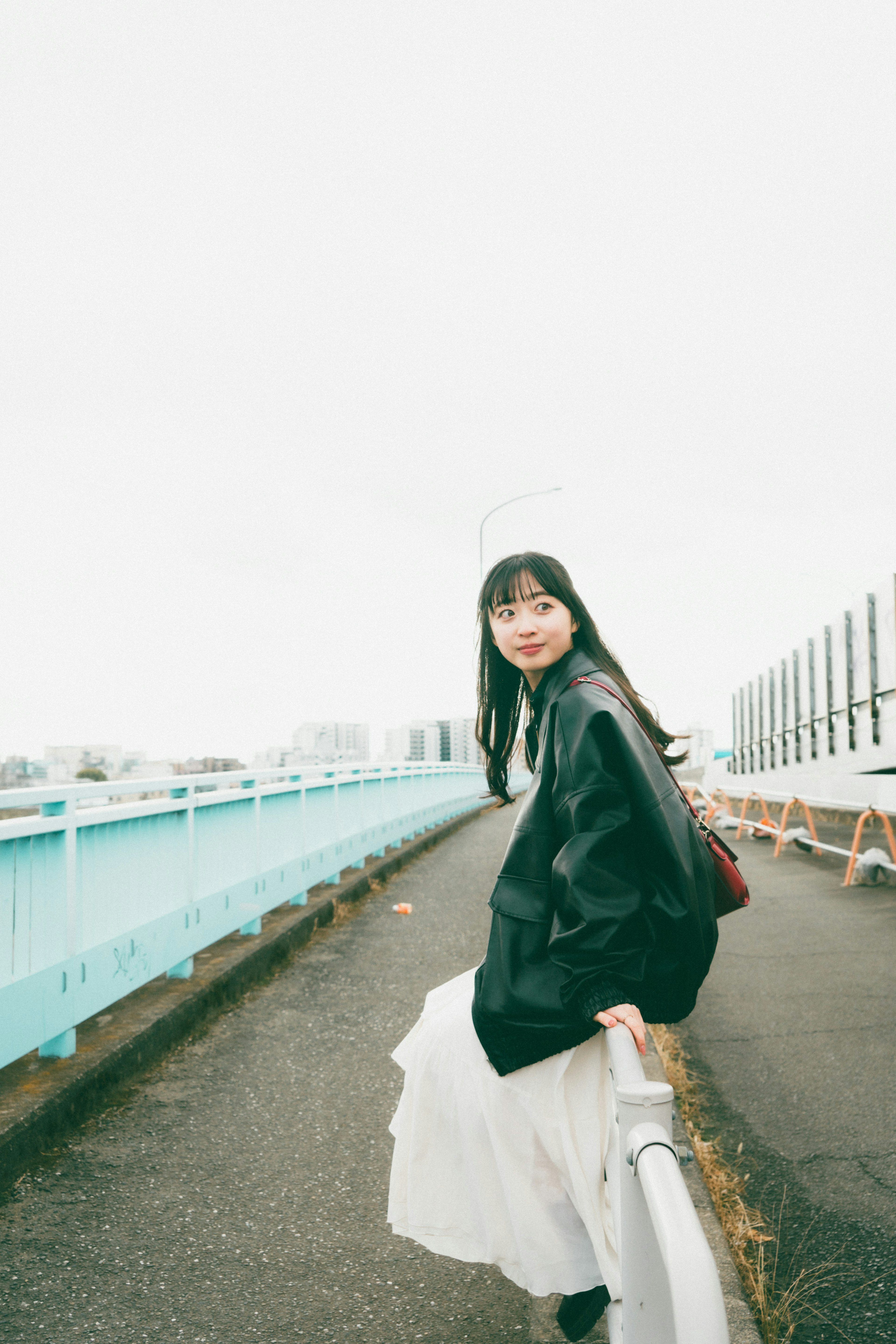 Eine Frau in einem weißen Rock und einer schwarzen Jacke, die auf einer blauen Brücke lächelt