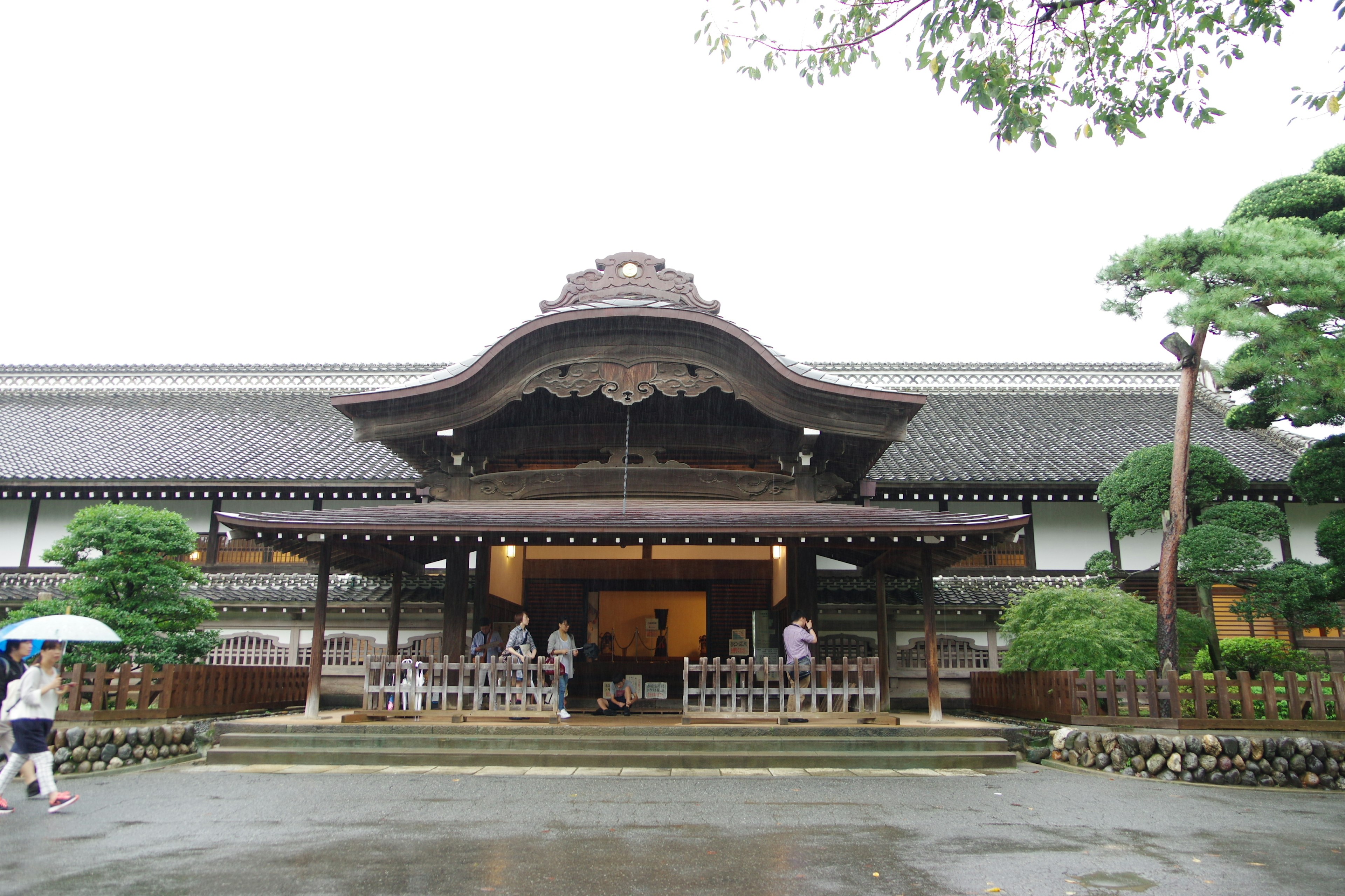 Traditional Japanese building facade in a rainy setting with green trees and stone pathway