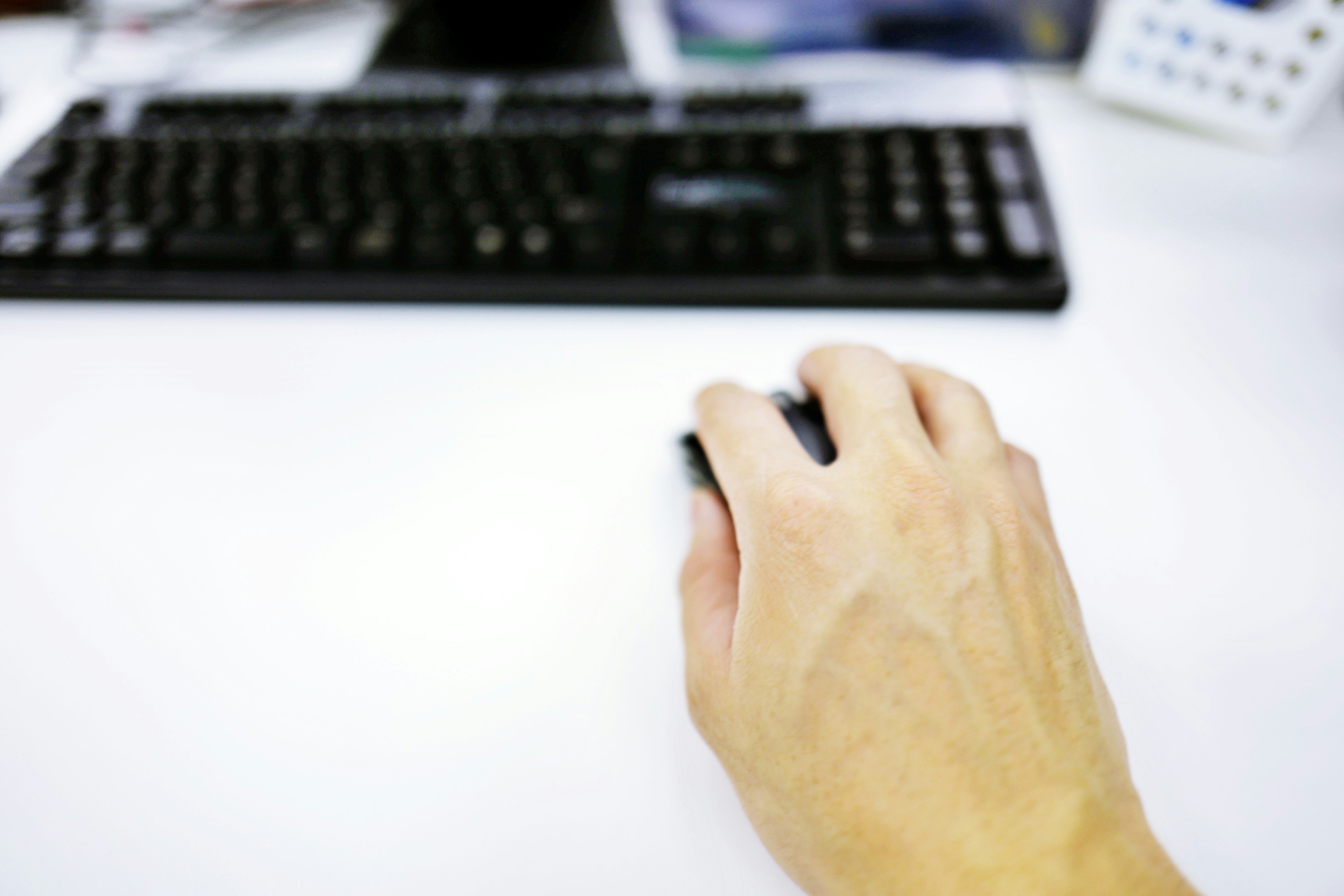 Mano usando un ratón sobre una mesa blanca con un teclado negro al fondo