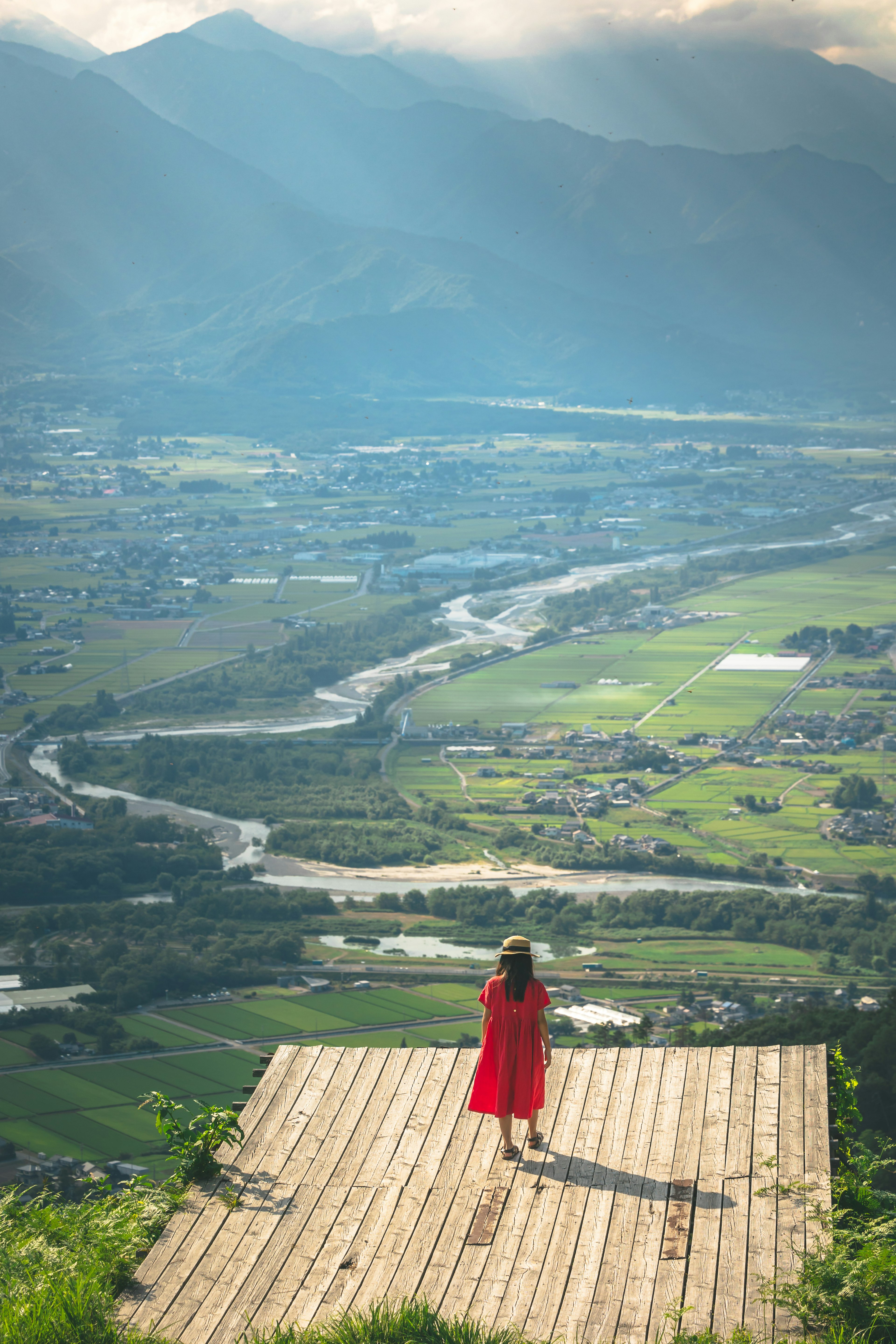 Eine Frau in einem roten Kleid steht auf einer Holzplattform mit Blick auf ein grünes Tal und einen Fluss