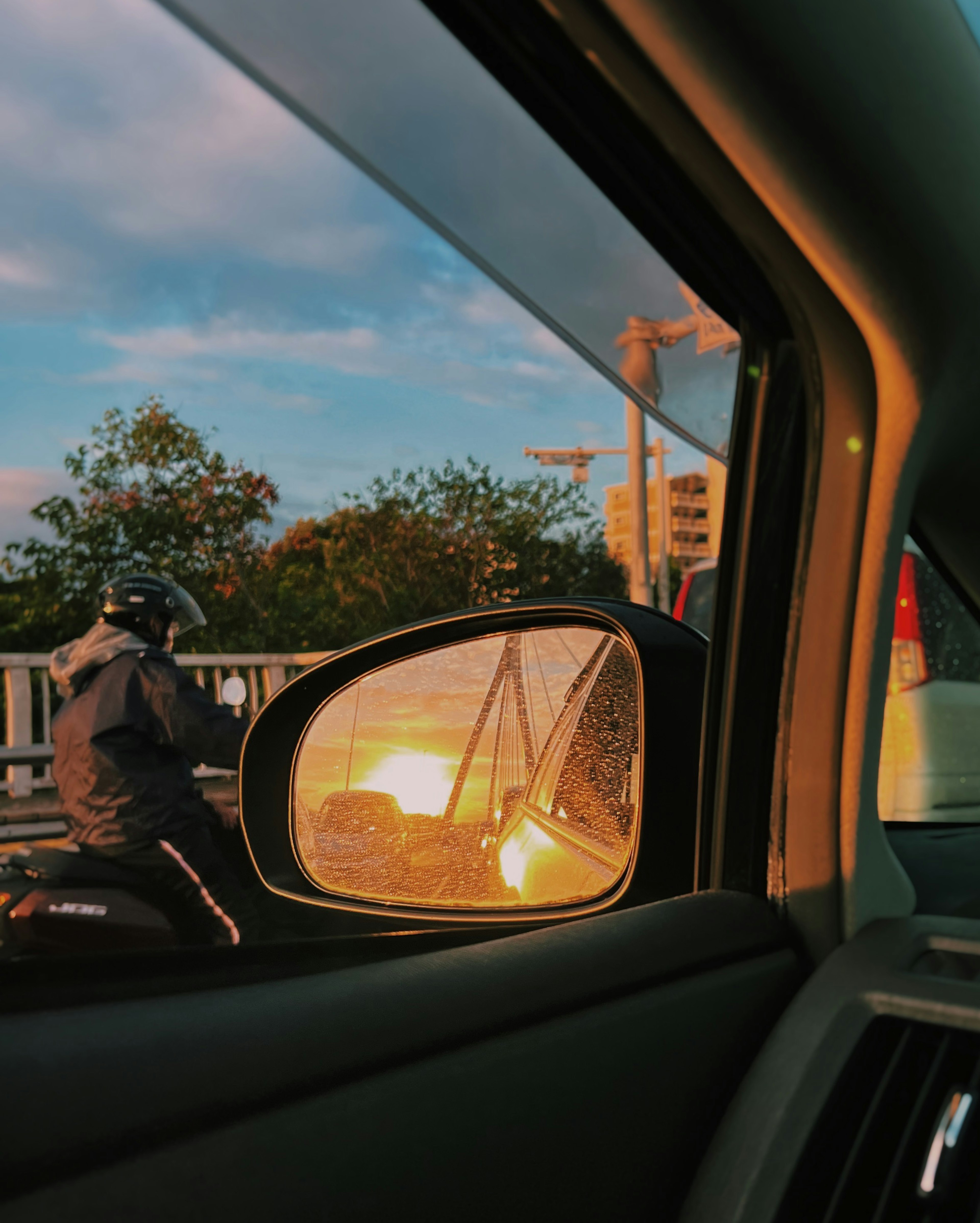 Refleksi matahari terbenam di cermin samping mobil dengan pengendara sepeda motor