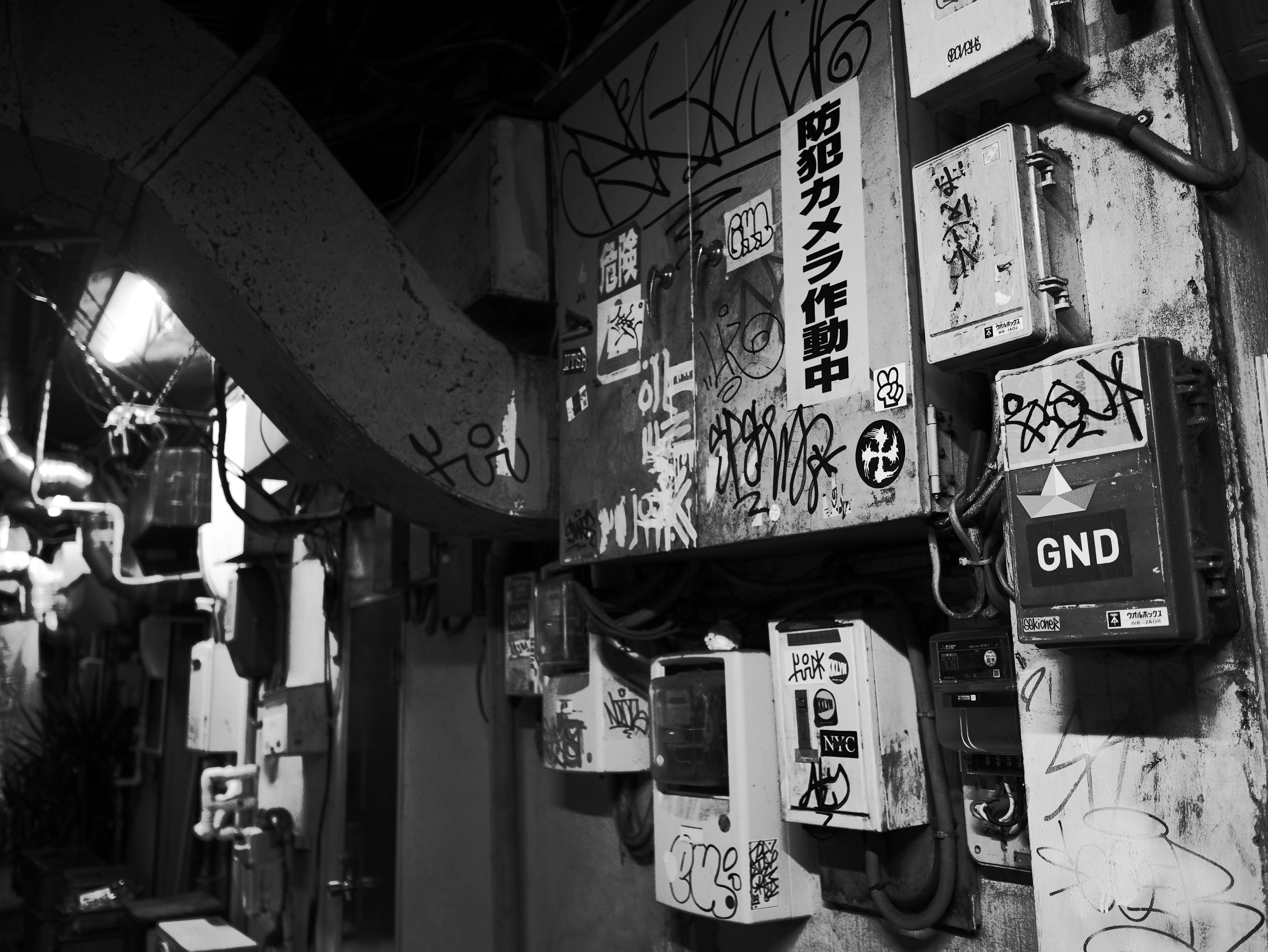 Photo en noir et blanc d'une ruelle étroite avec des cabines téléphoniques anciennes et des équipements sur un mur couvert de graffitis