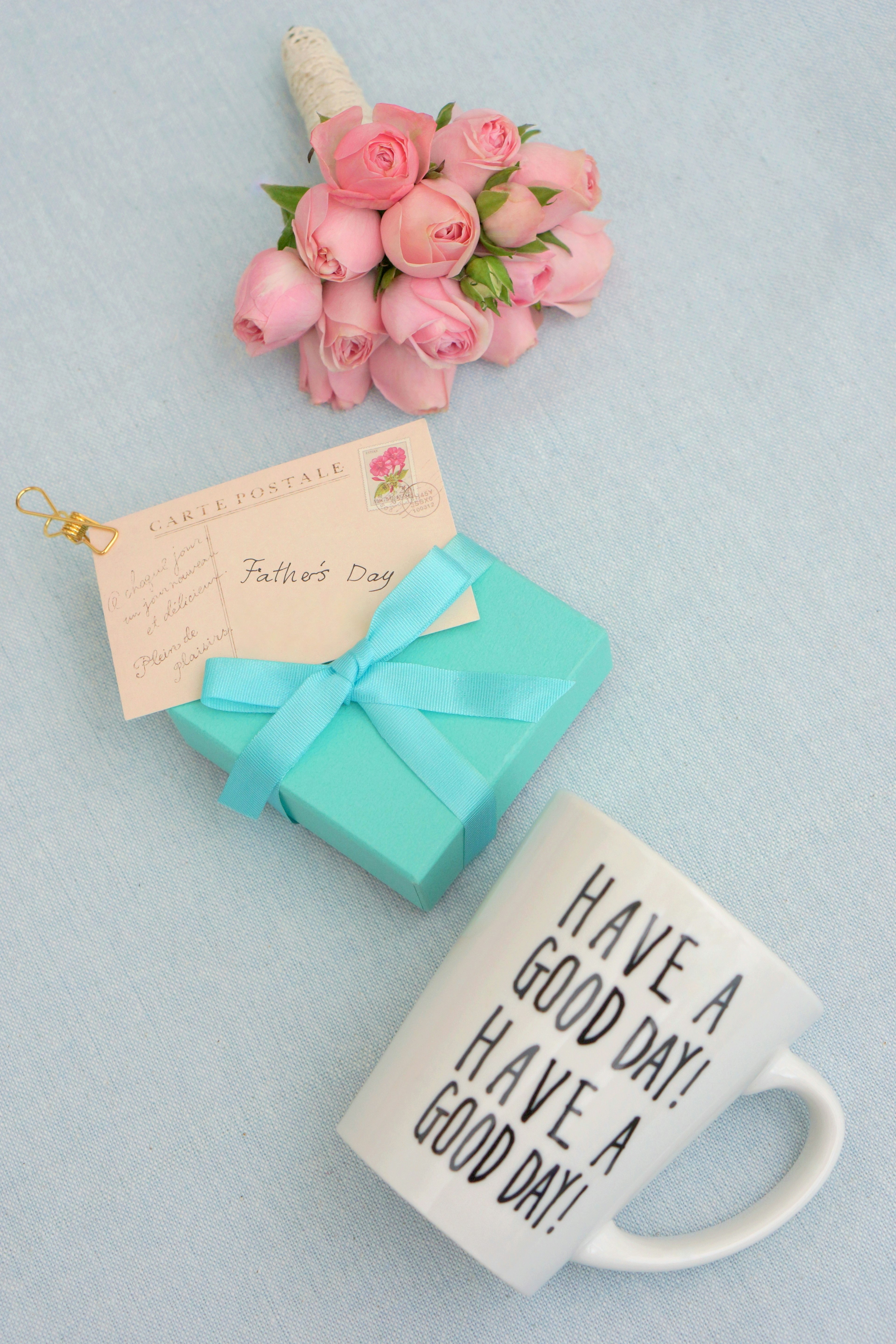 Un bouquet de fleurs roses, un cadeau enveloppé en turquoise avec une étiquette, et une tasse blanche avec le message Avoir une bonne journée sur un fond bleu