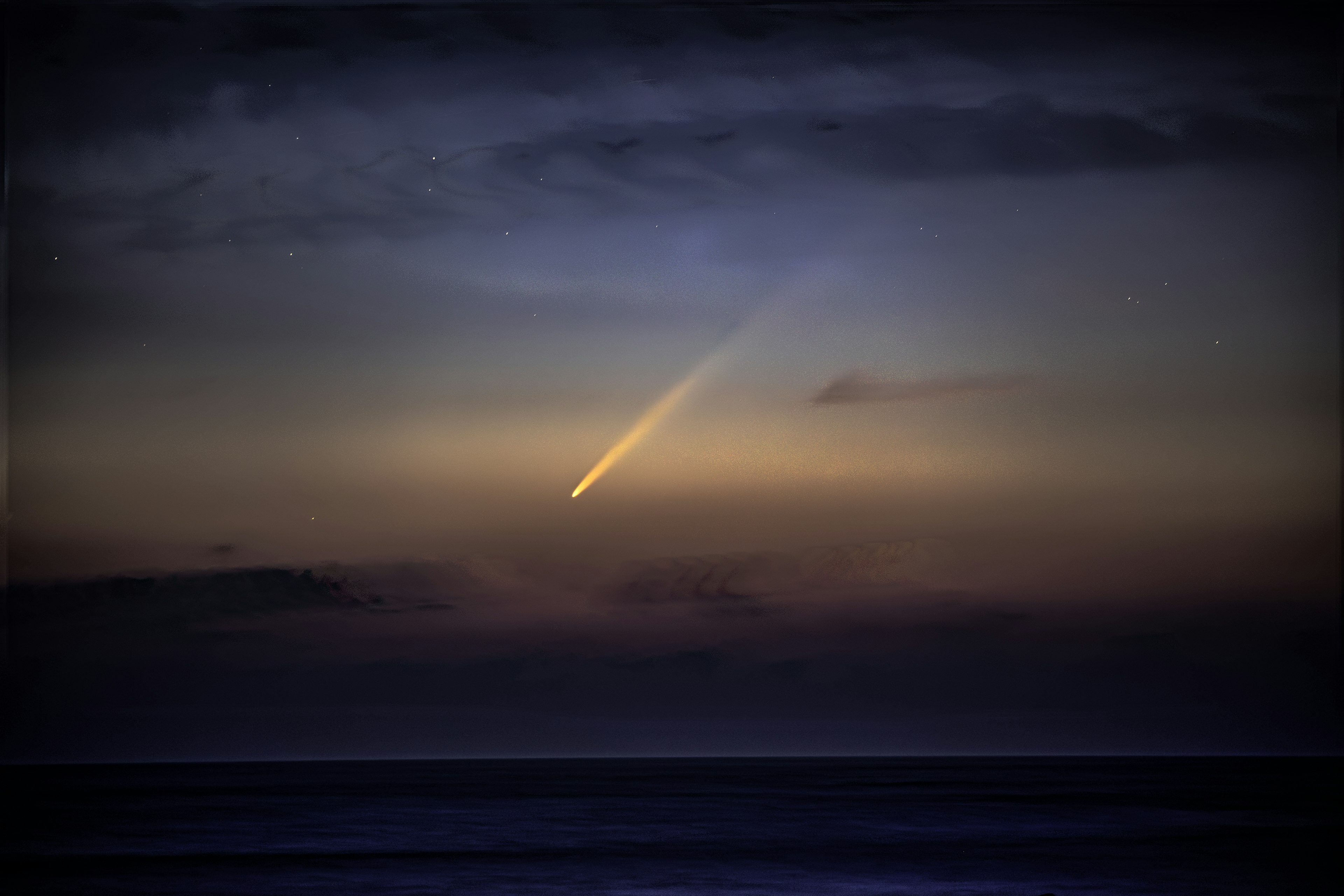 A comet's tail shining over the ocean at dusk with stars in the sky