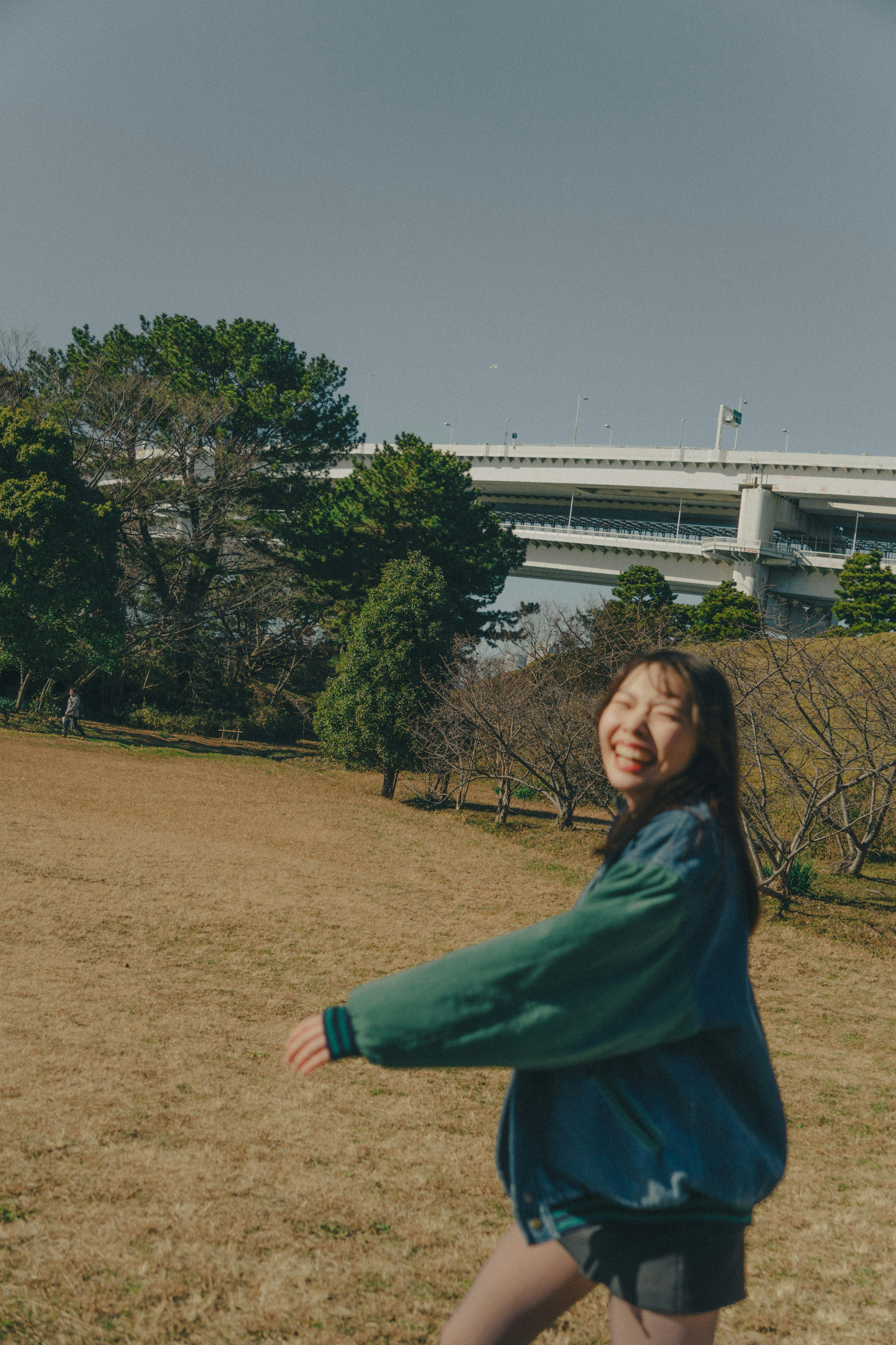 公園裡微笑著走的女性 穿著綠色夾克 背景有高架橋和樹木