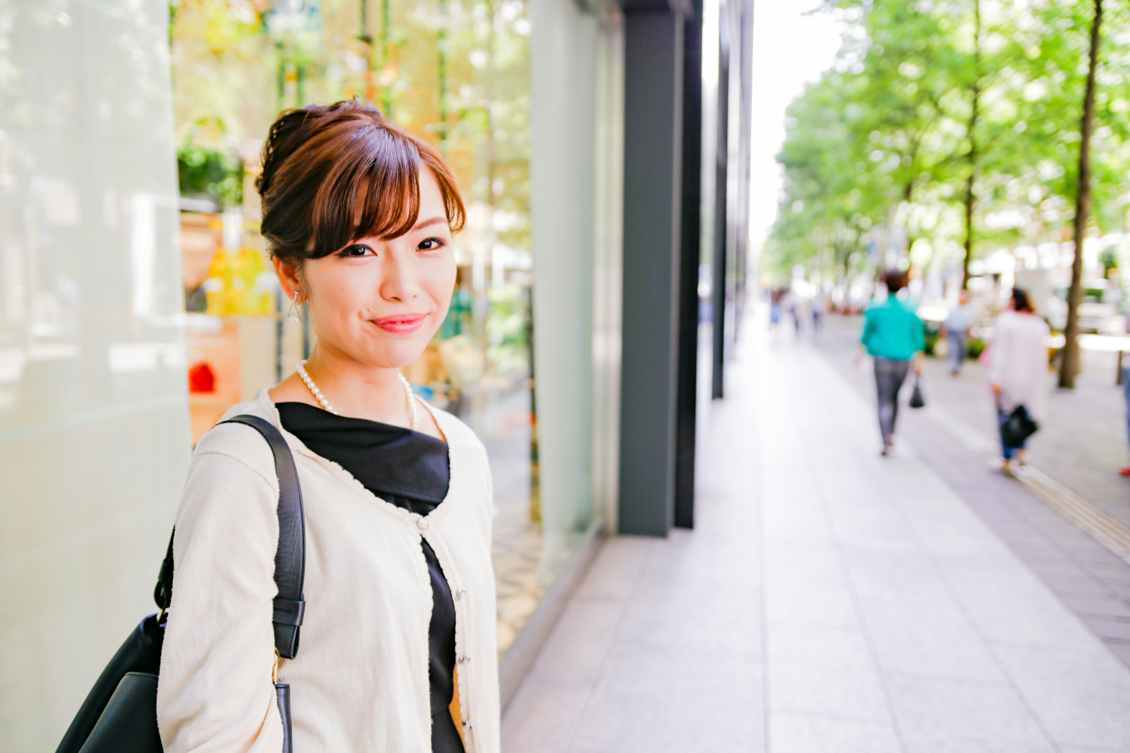 A smiling woman standing on a city sidewalk wearing a light-colored outfit with a black scarf green trees and pedestrians in the background