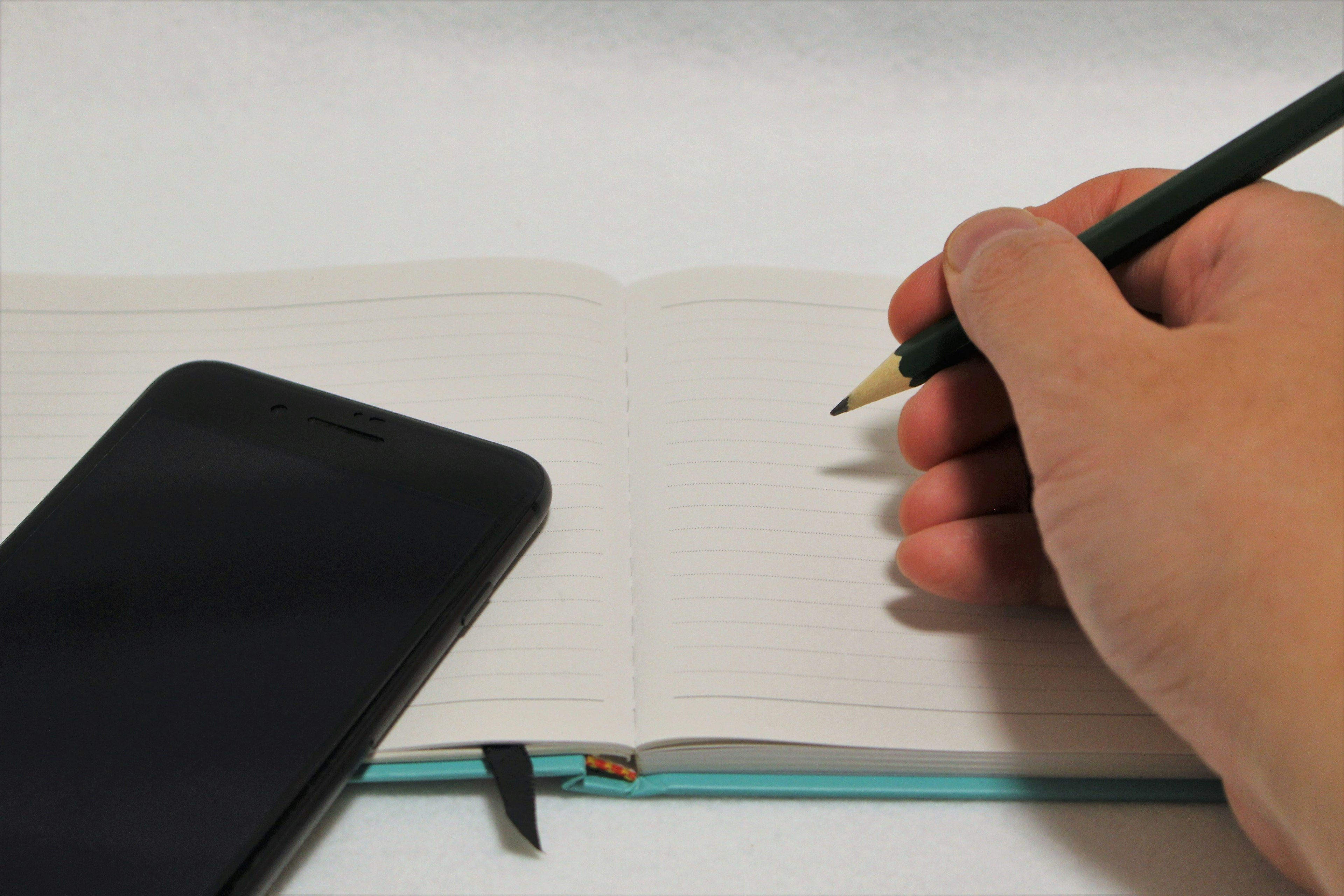 A hand holding a pencil writing in a notebook with a smartphone beside it