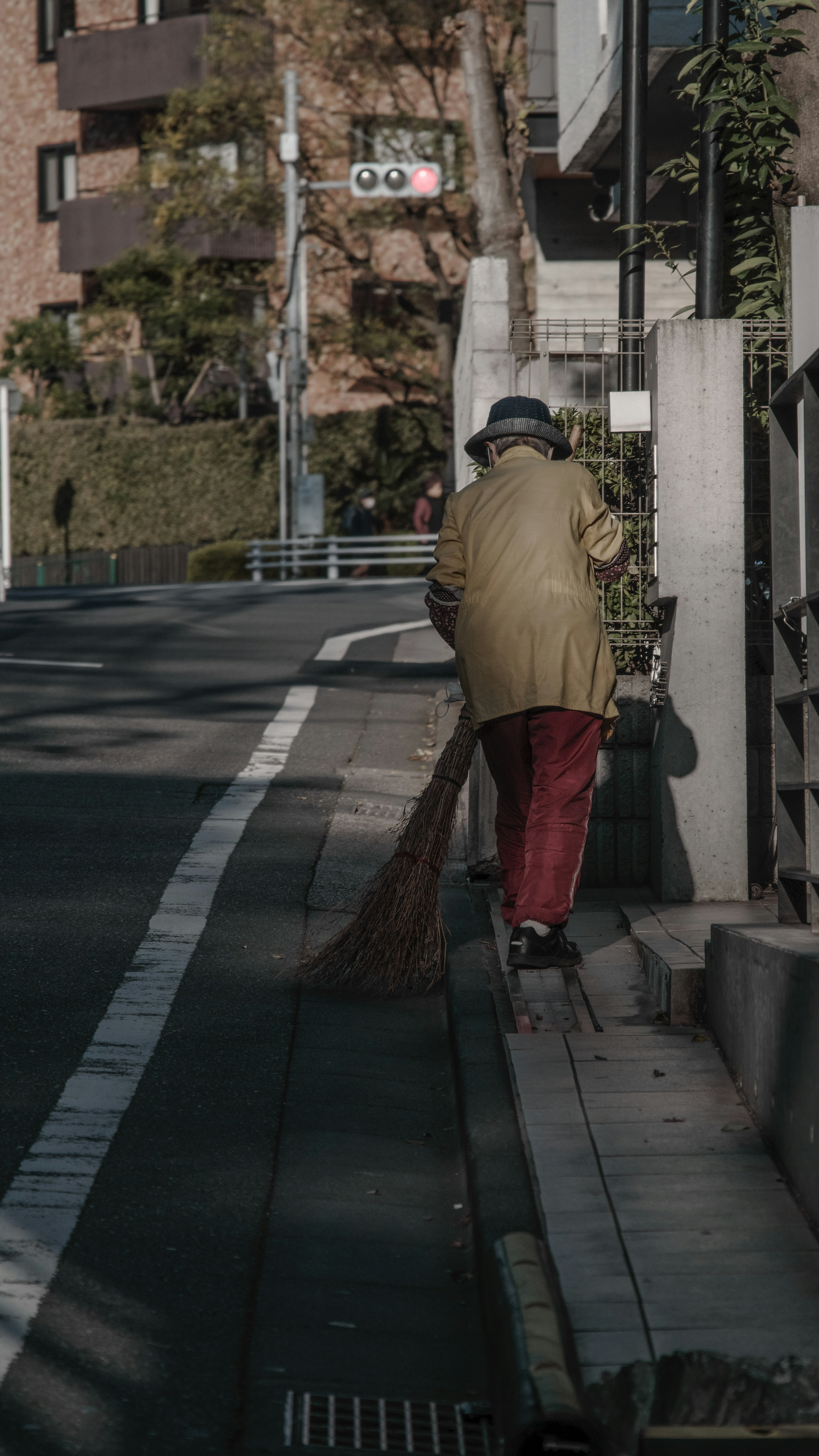 ชายชรา sweeping ถนนสวมเสื้อโค้ทสีน้ำตาลและกางเกงแดง