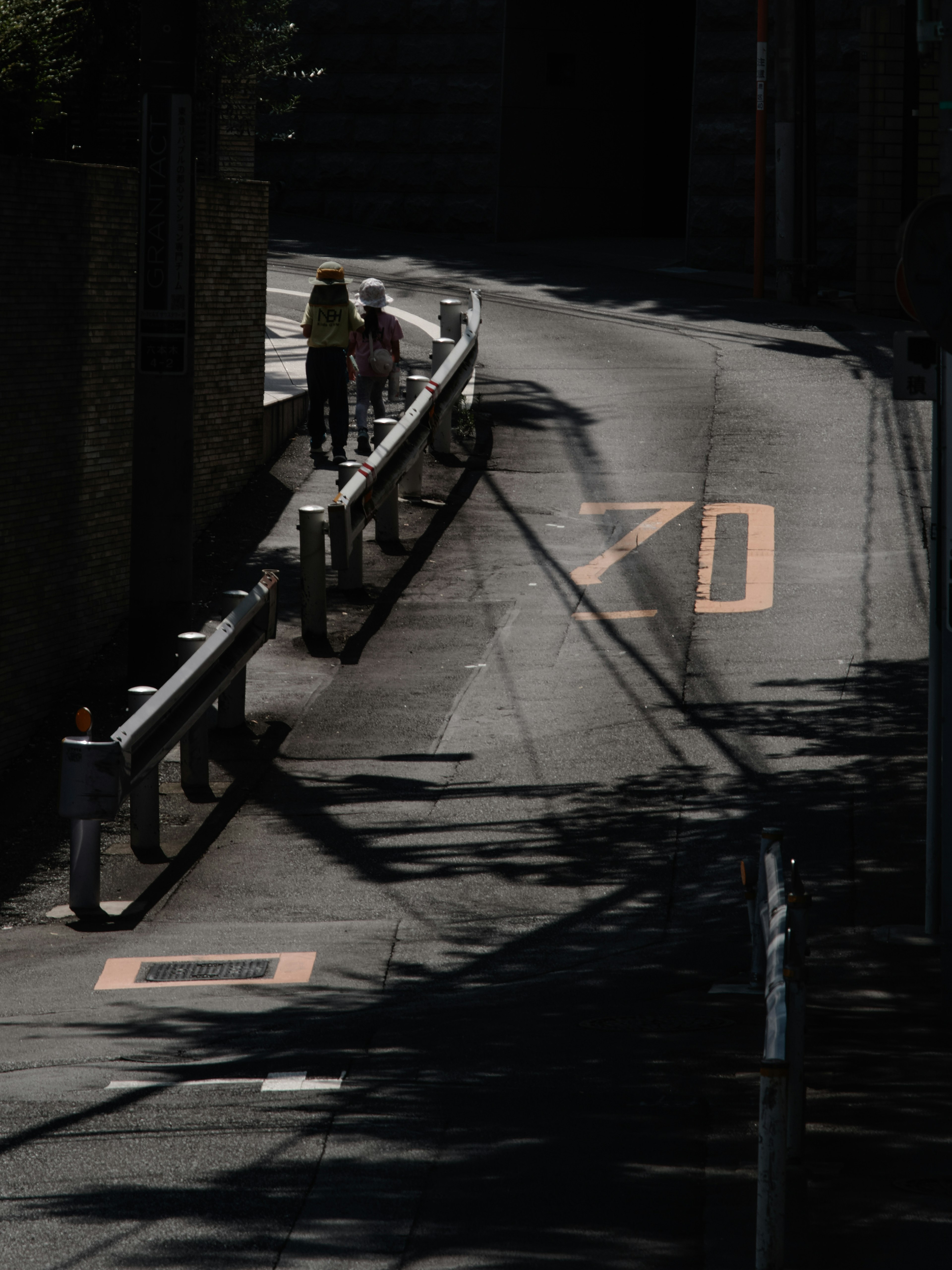 Image of a sloped road with people walking Shadows cast by a guardrail and road markings are visible