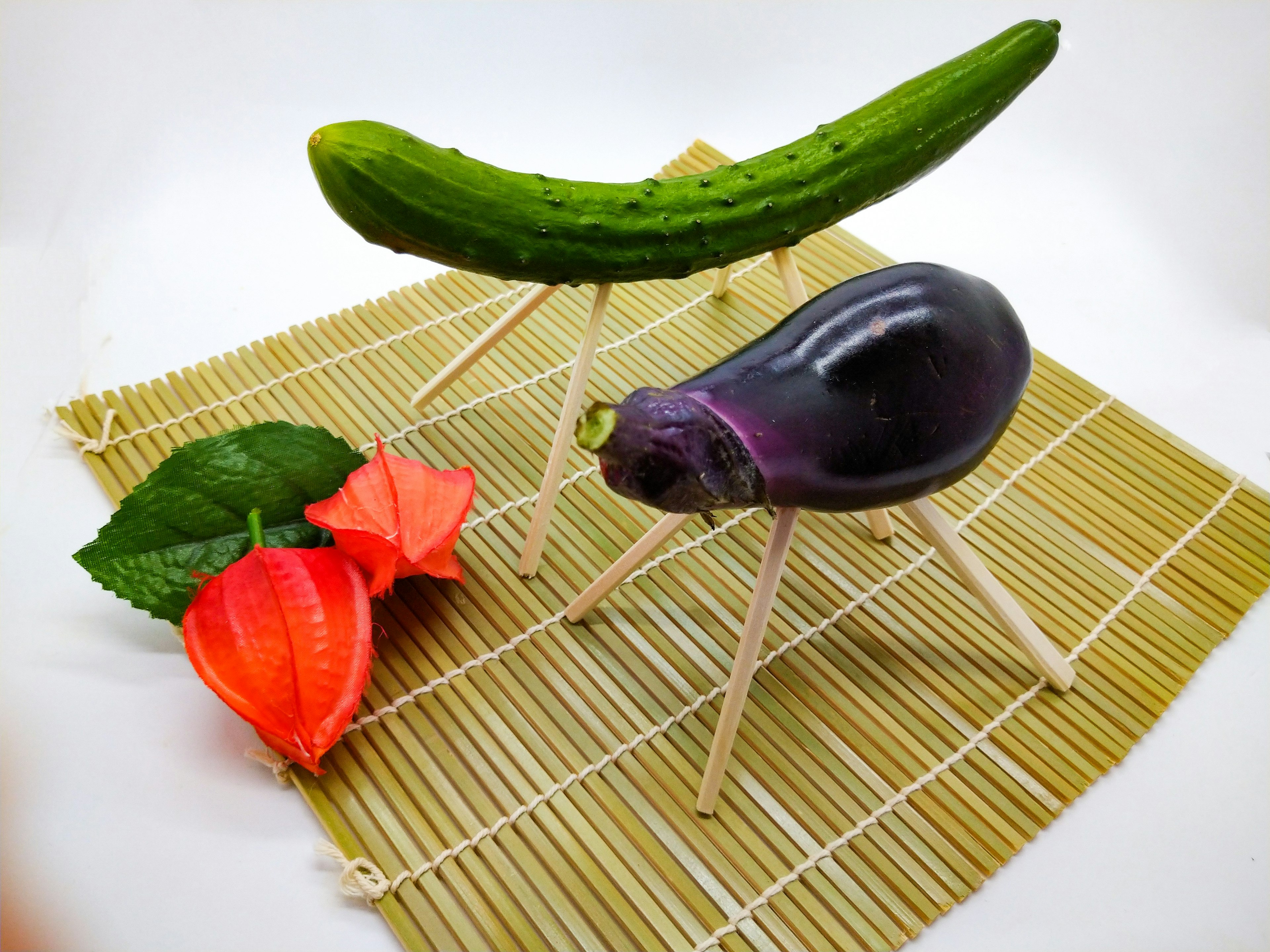 Creative vegetable art featuring a cucumber and eggplant on a bamboo mat with orange flowers