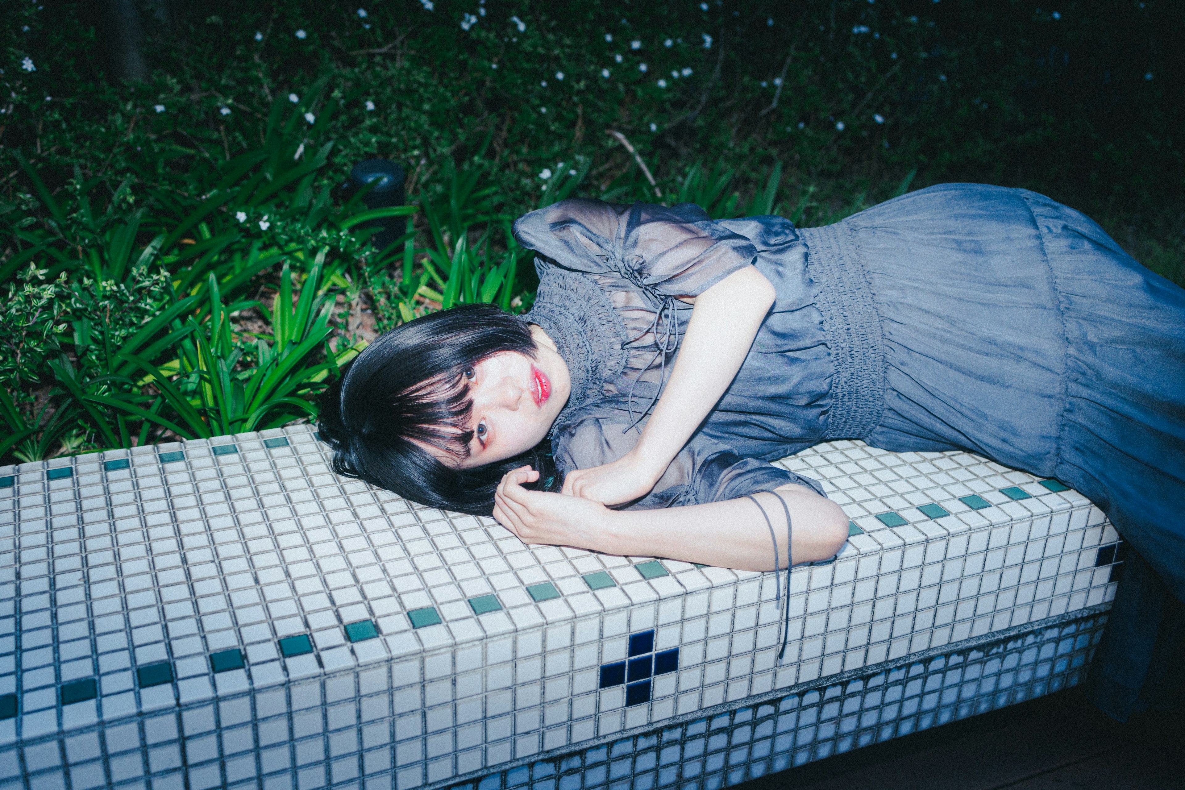 A woman lying on a tiled bench in a dark setting wearing a gray dress and black hair