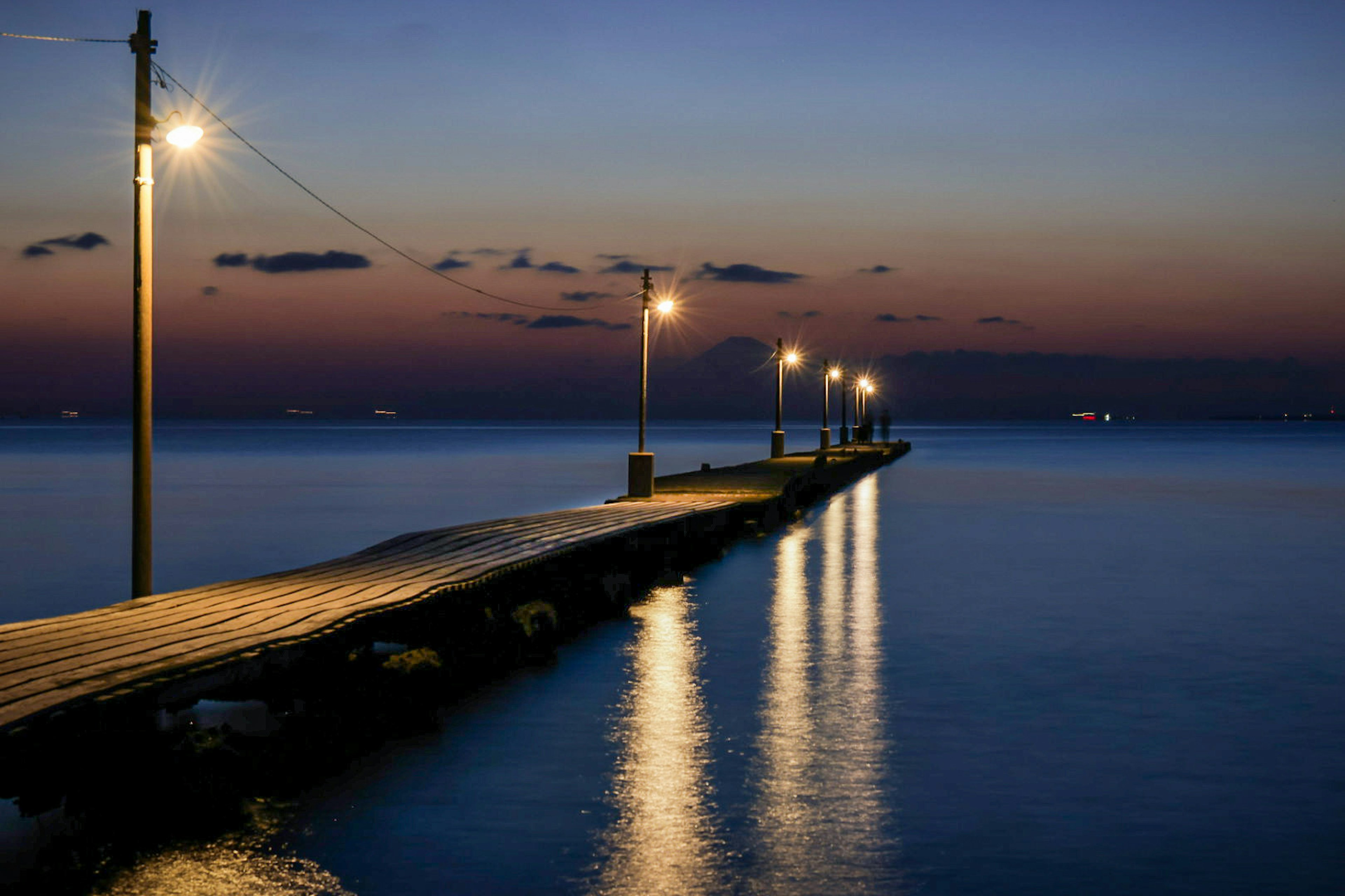 Imagen de un muelle junto al mar al anochecer luces iluminando el camino reflejándose en el agua tranquila