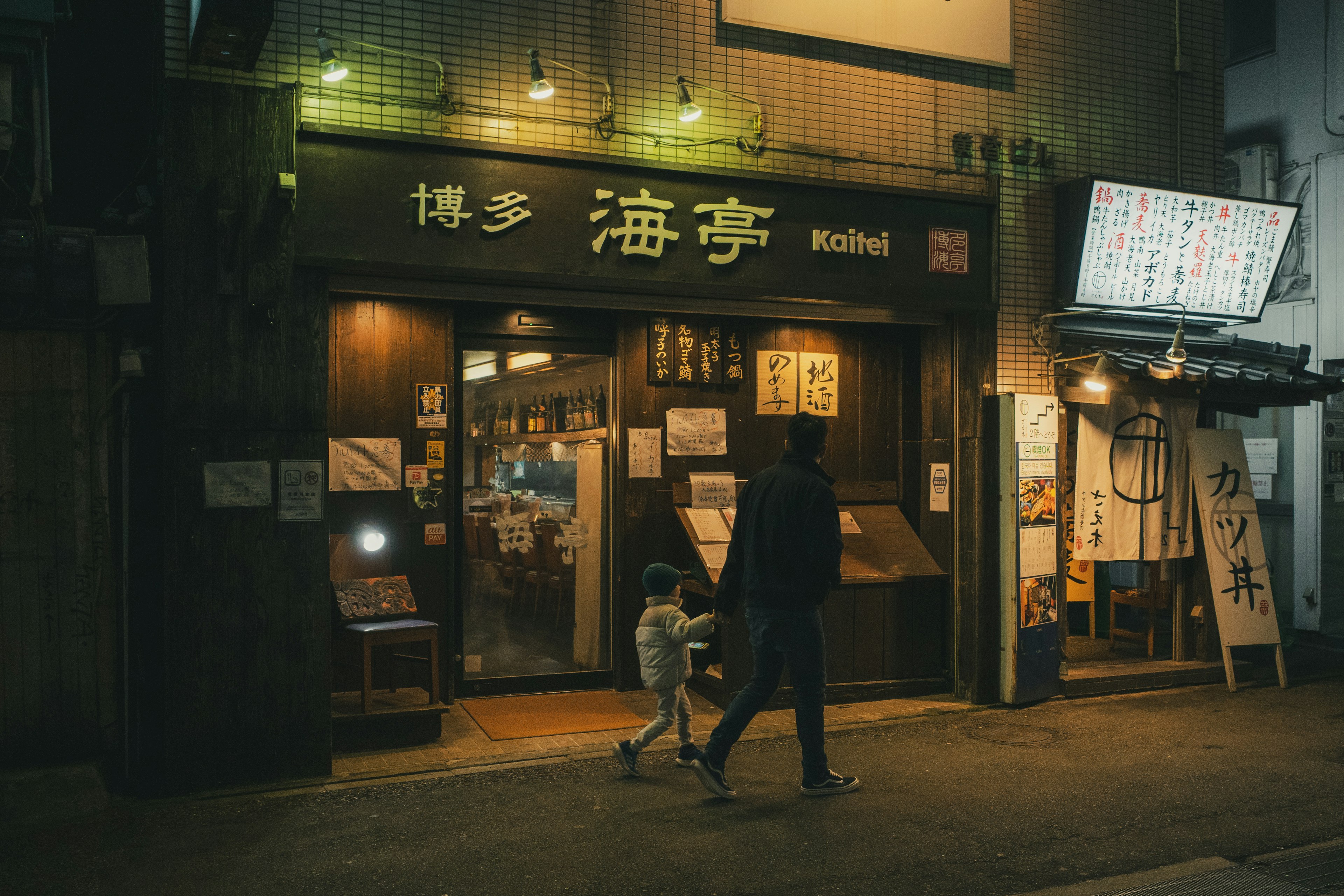 Un padre y un hijo caminando por las calles por la noche frente a un restaurante con un letrero que dice '海亭'