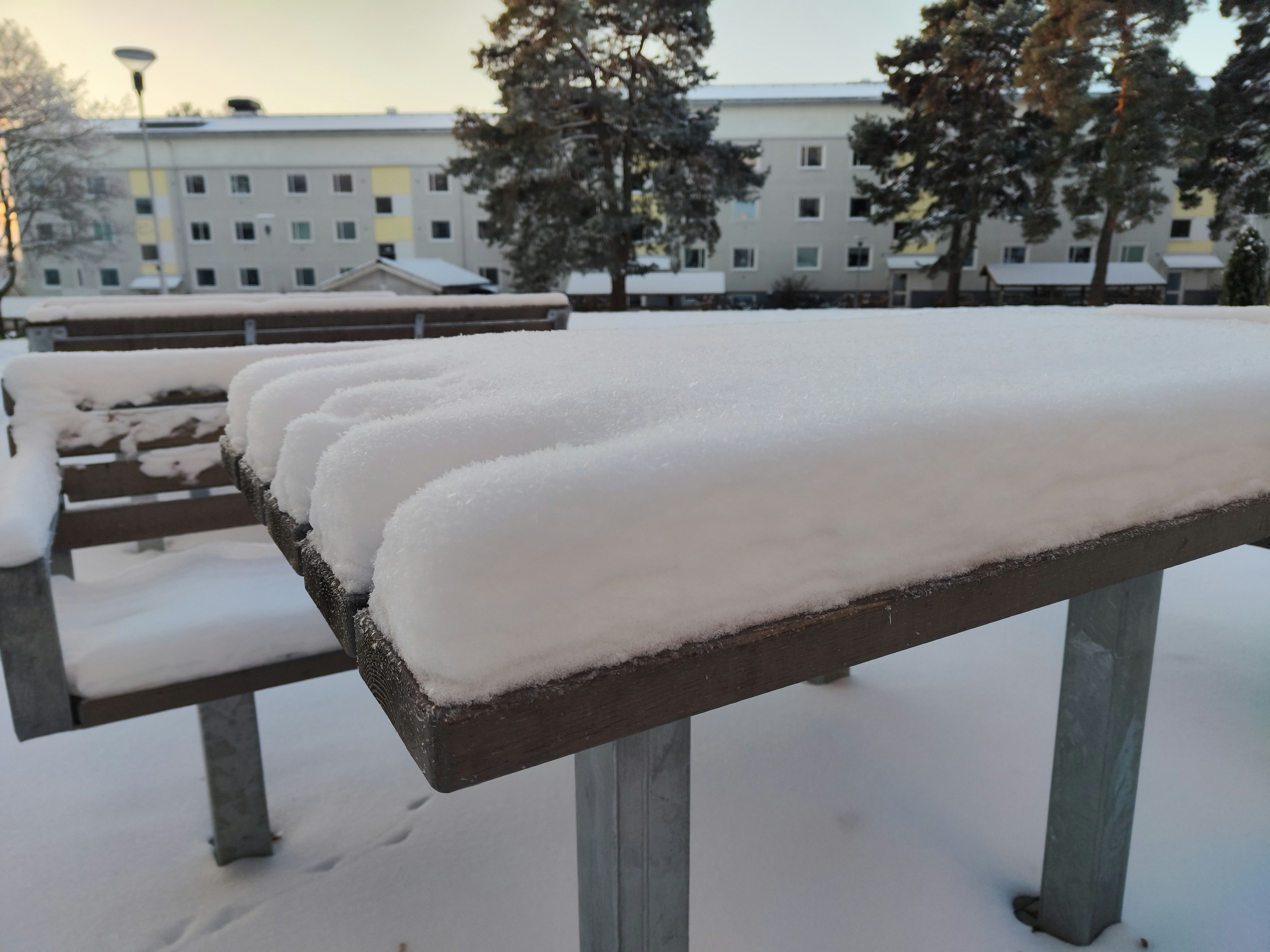 Scena invernale con una panchina coperta di neve e edifici sullo sfondo