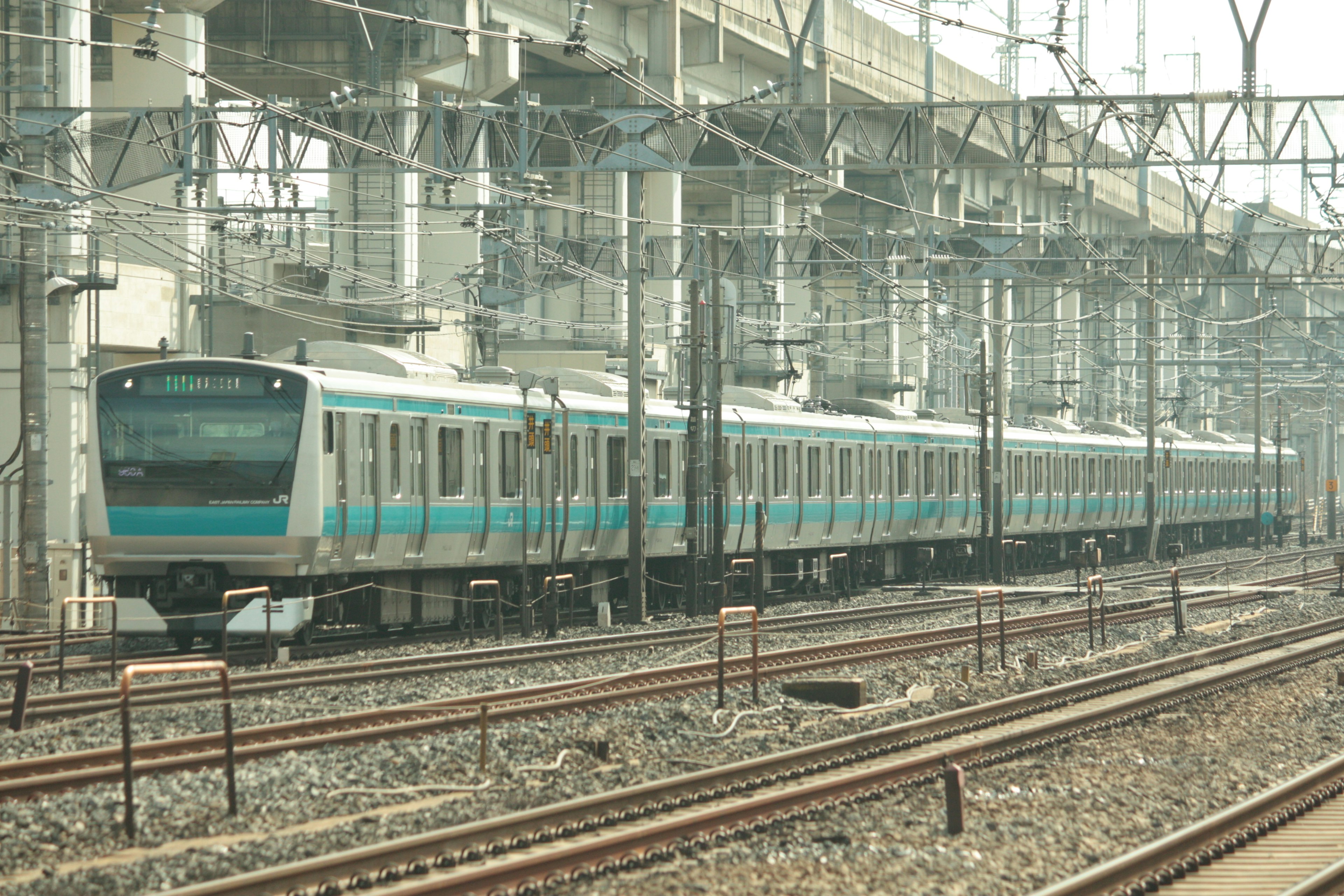 A blue train traveling along the tracks with overhead wires and structures in the background