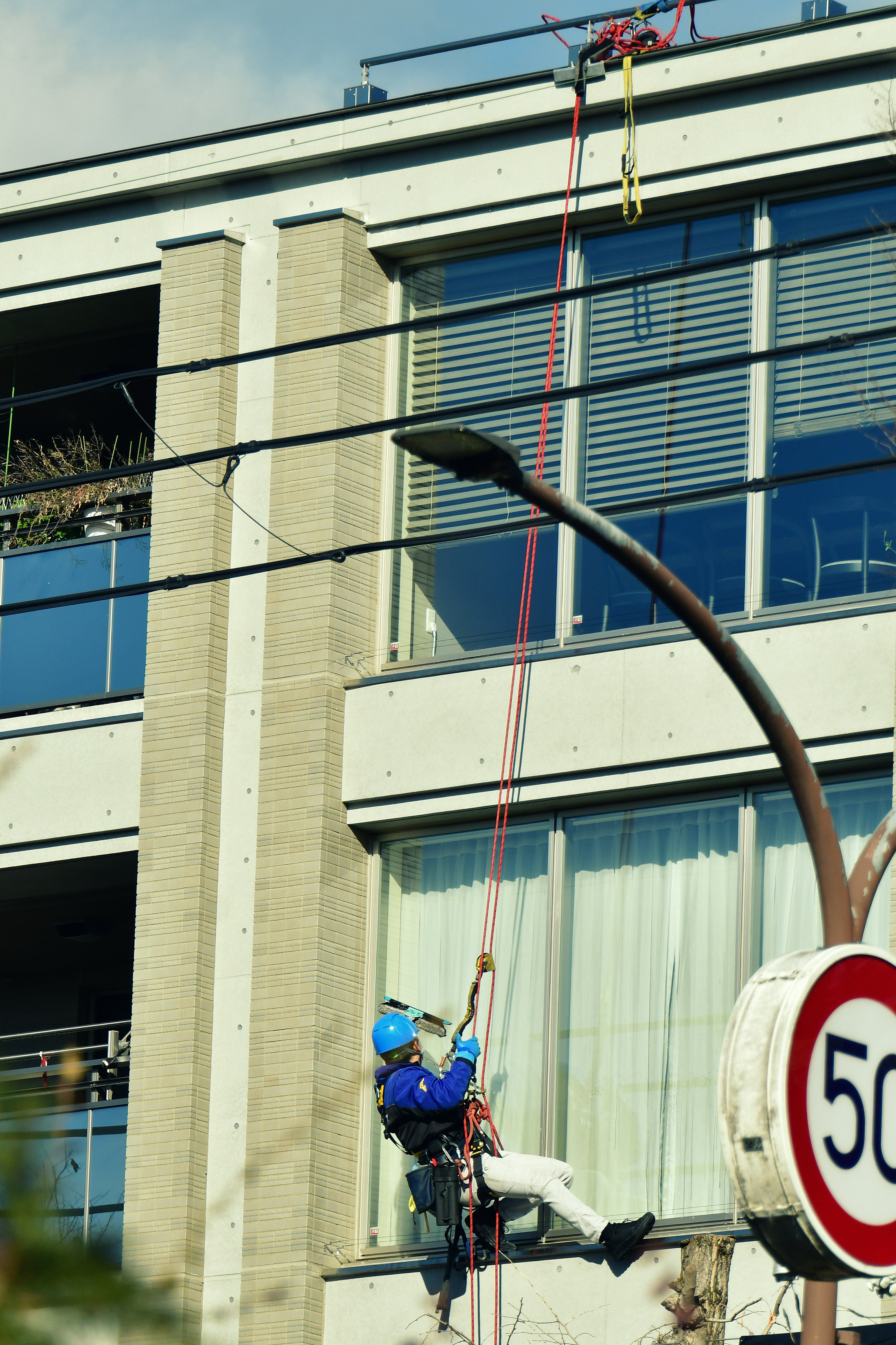 Trabajador usando cuerdas fuera de un edificio con un casco azul y un letrero de límite de velocidad 50 visible