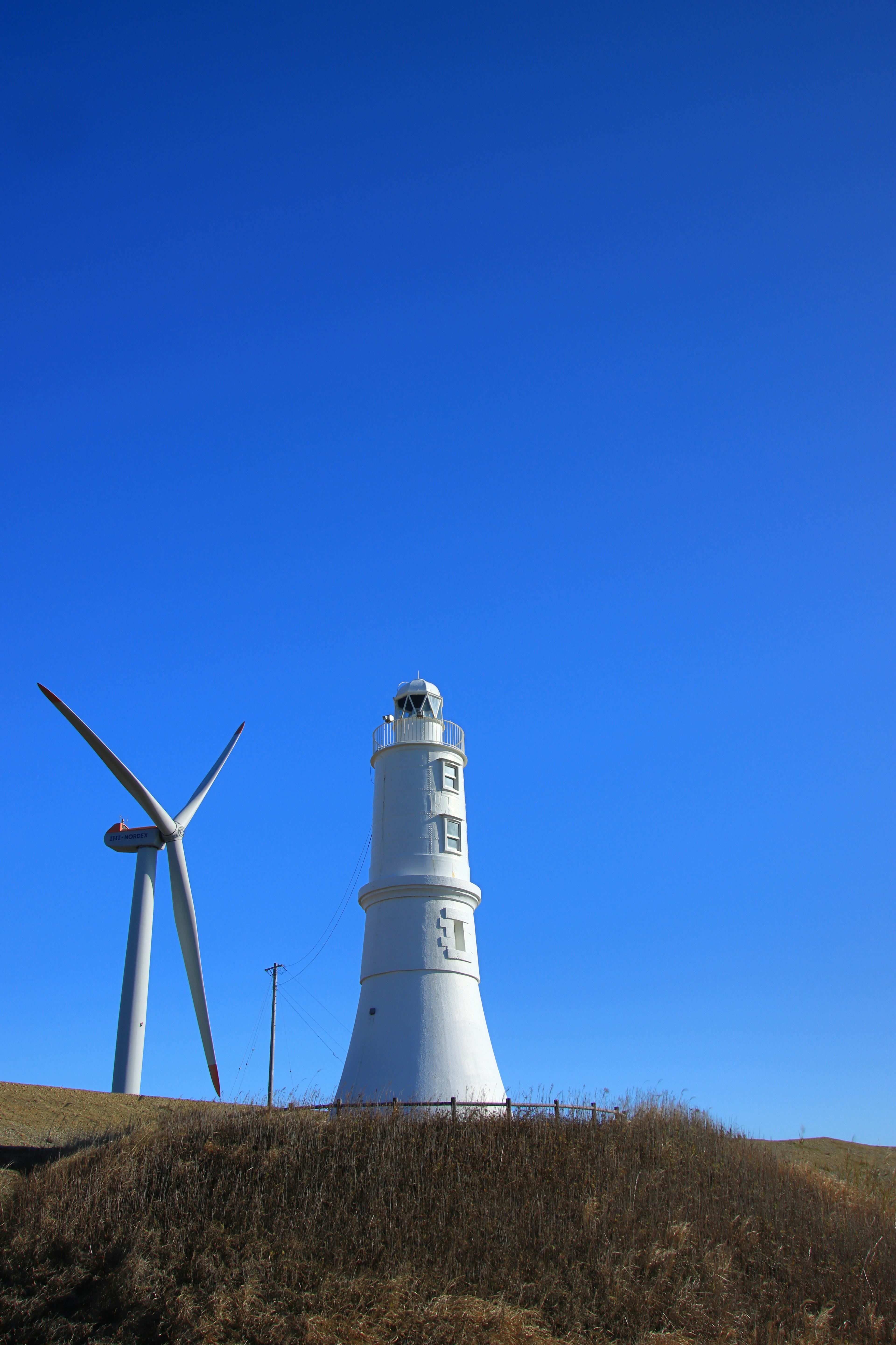 Faro blanco junto a una turbina eólica contra un cielo azul claro