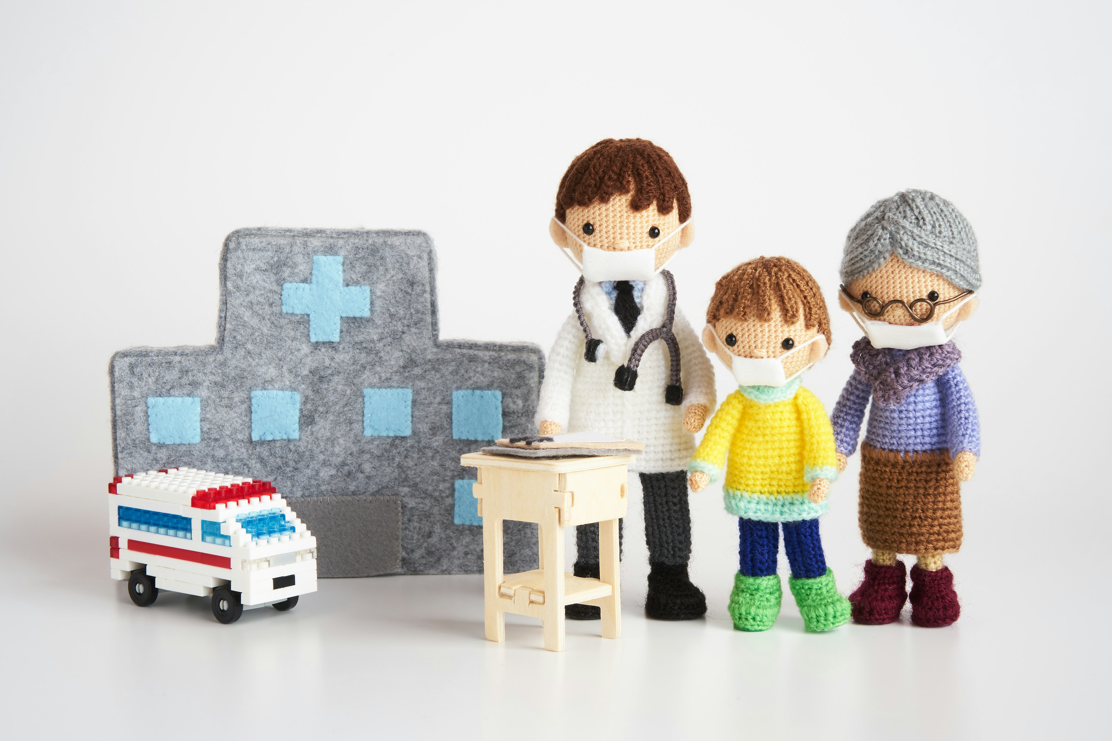 A scene featuring plush toys and props with a doctor, child, and grandmother wearing masks in front of a hospital backdrop and a miniature ambulance