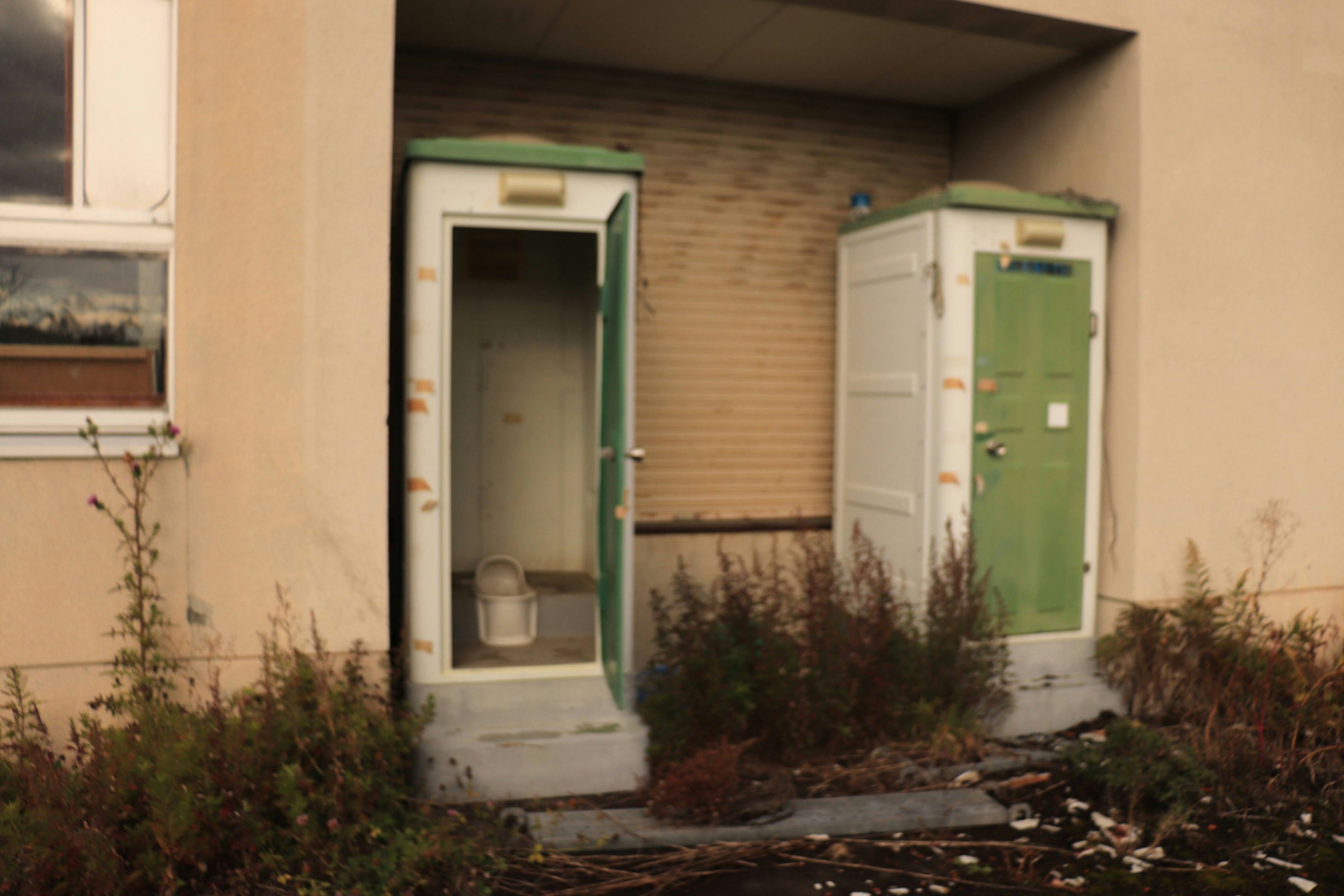 Two old public toilets with green doors outside an abandoned building surrounded by overgrown grass