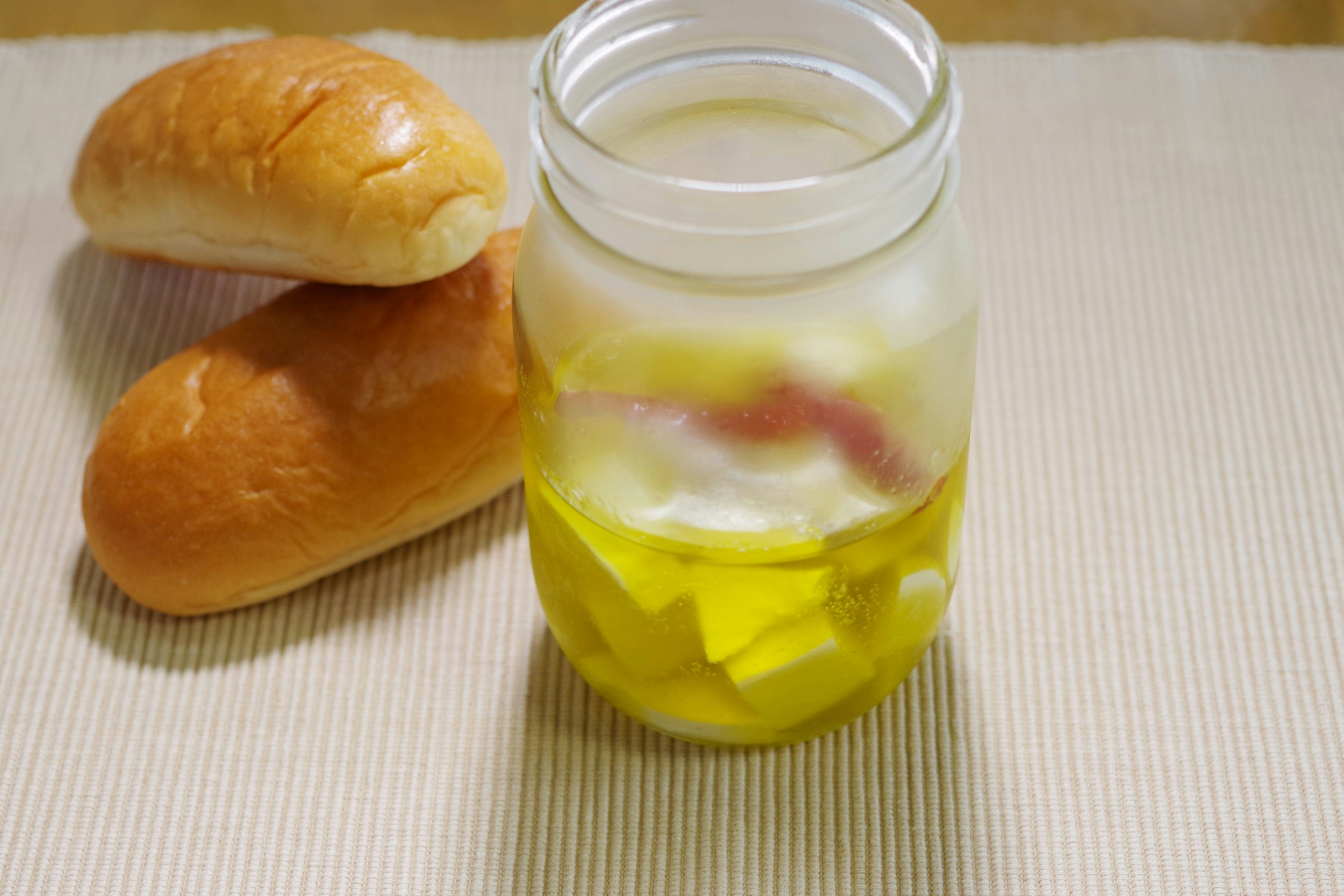 Jar filled with olive oil and chopped ingredients next to bread rolls