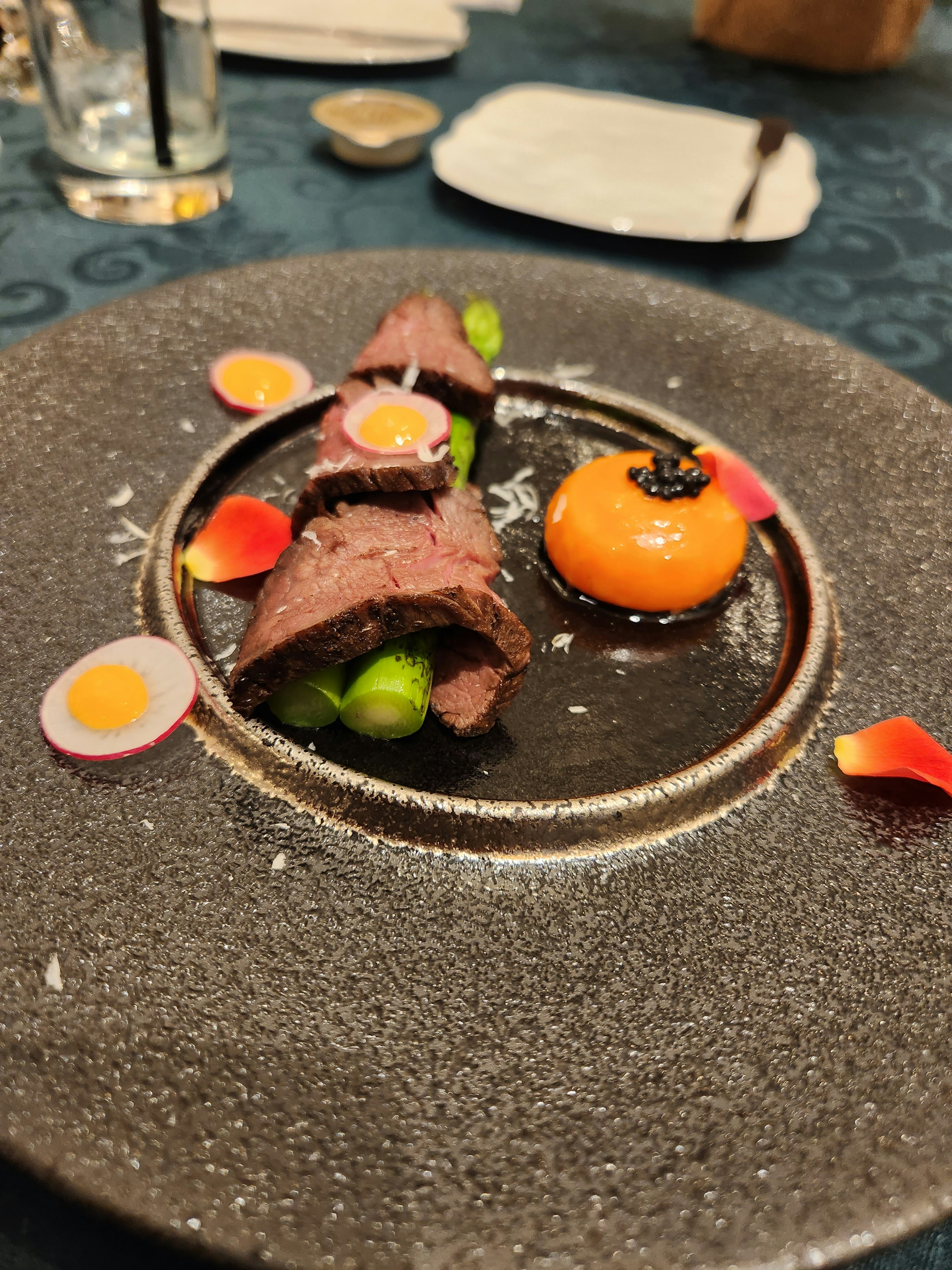 An elegant dish presented on a black plate featuring sliced meat with colorful garnishes including an orange sphere and assorted vegetables