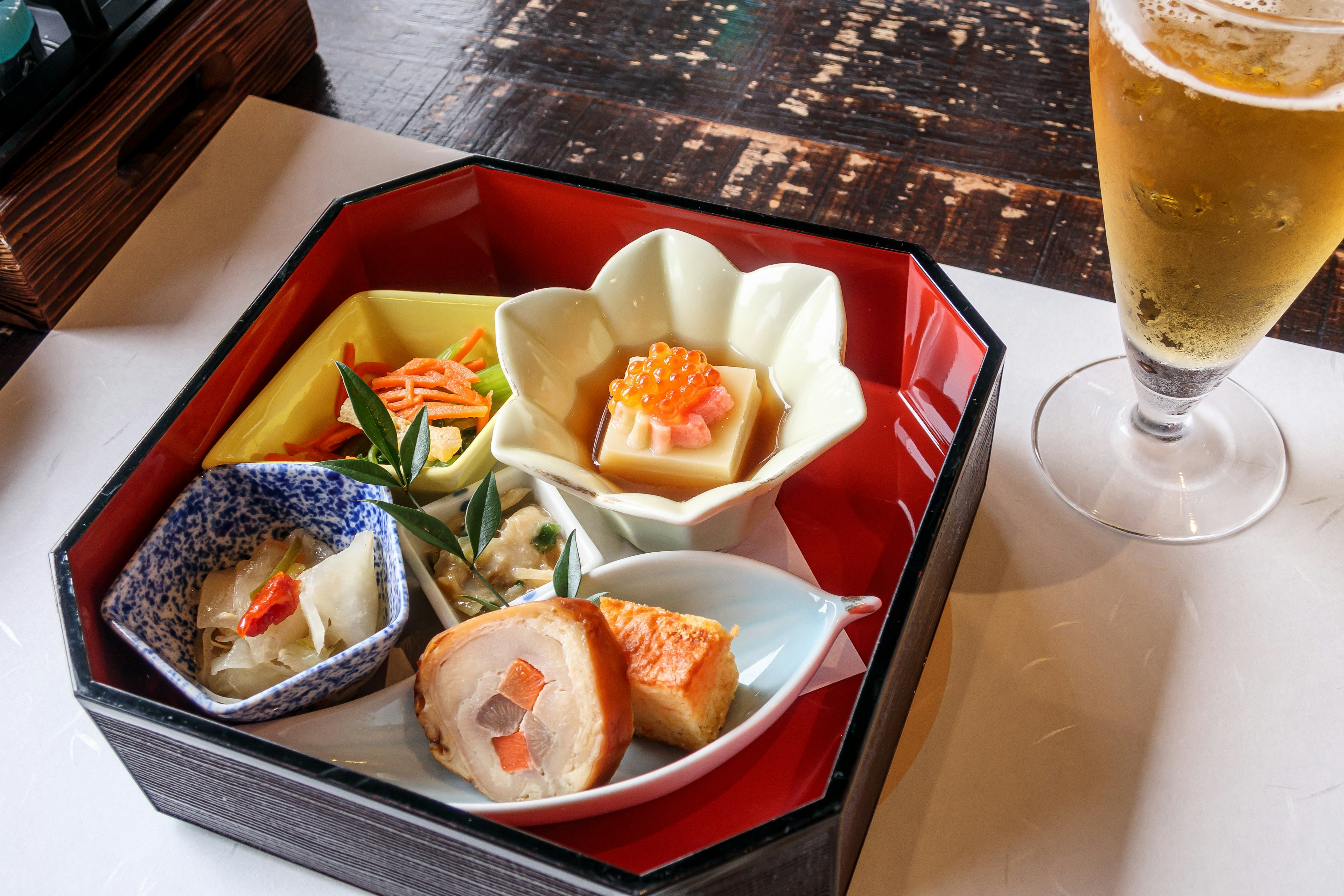 A beautifully arranged bento box with colorful Japanese dishes and a glass of beer