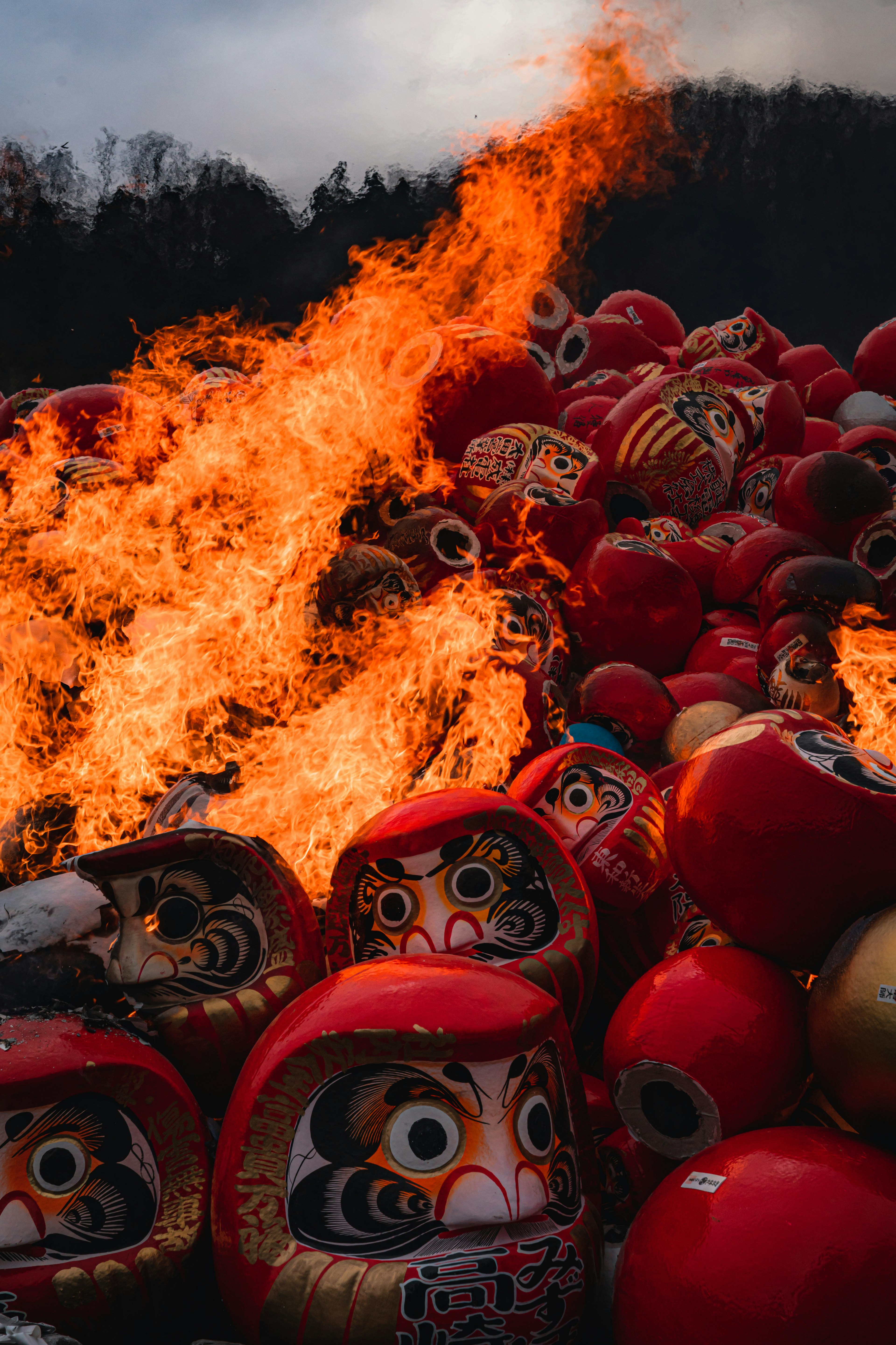 A pile of Daruma dolls engulfed in flames with a dark sky