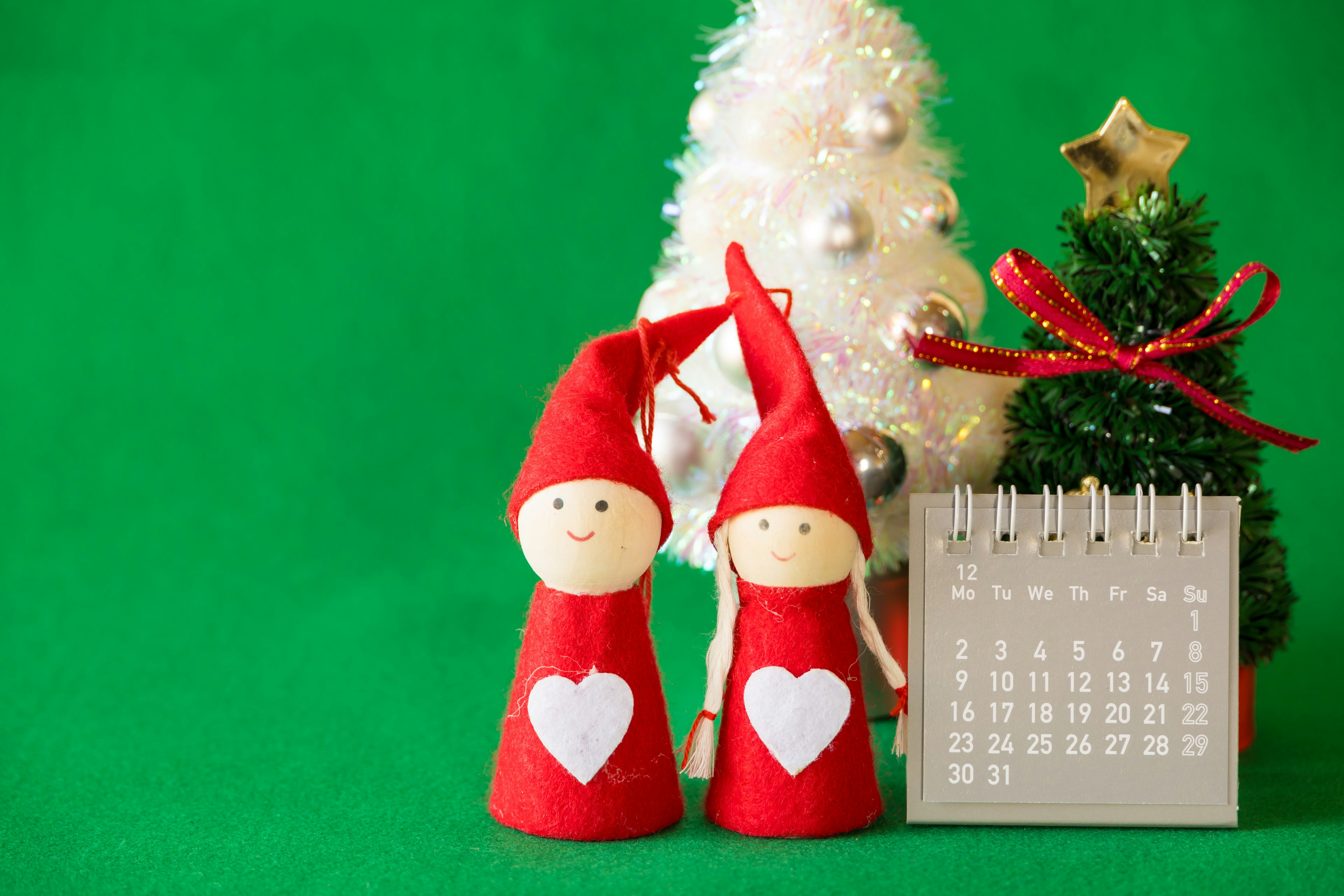 Two dolls in red hats with heart designs stand beside a small Christmas tree and a calendar on a green background