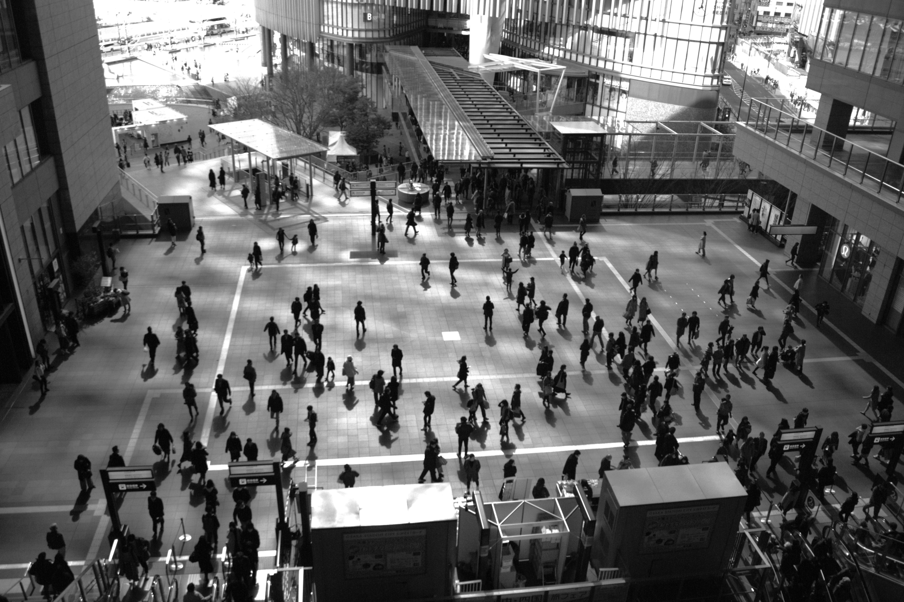 Black and white image of people walking in a plaza surrounded by tall buildings