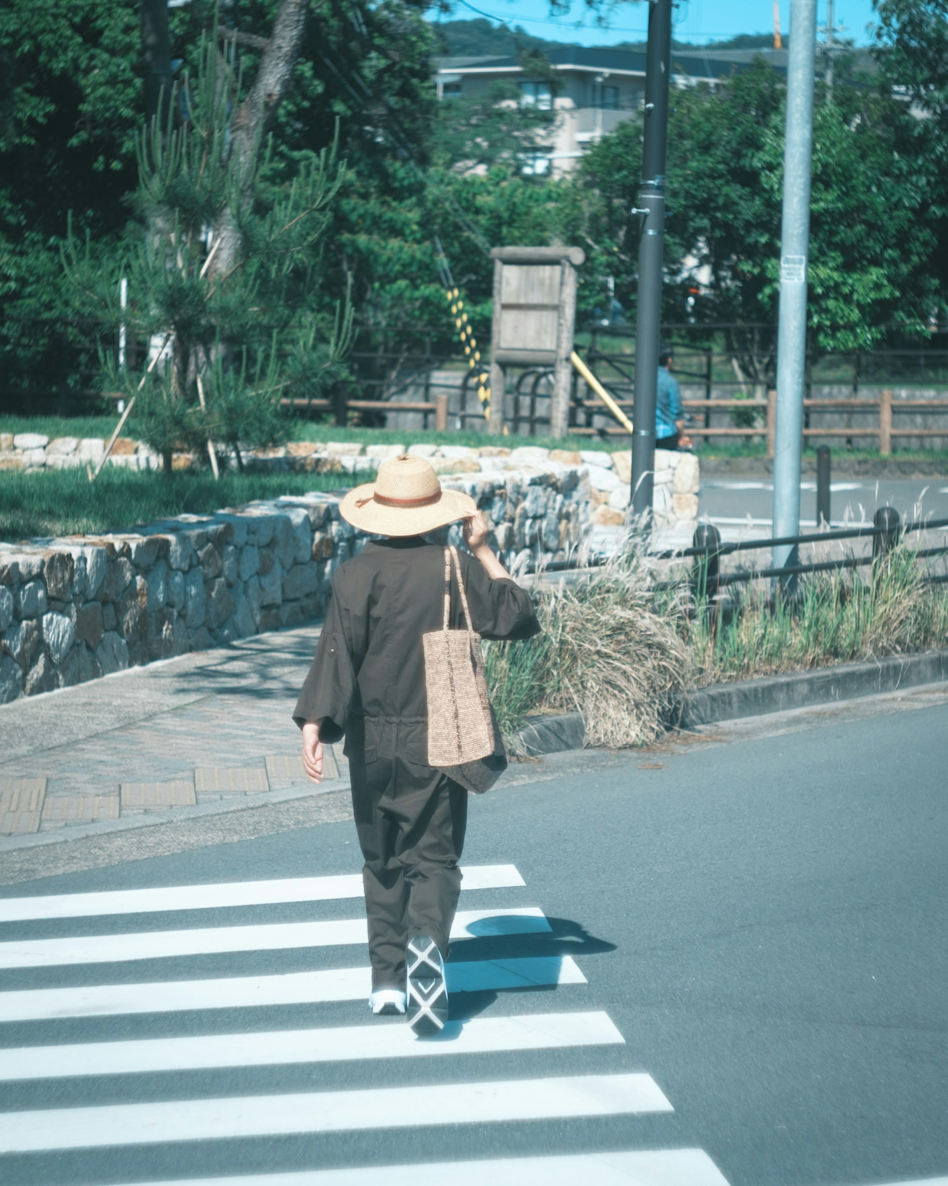 Person wearing a hat walking across a crosswalk with green trees and a stone wall in the background