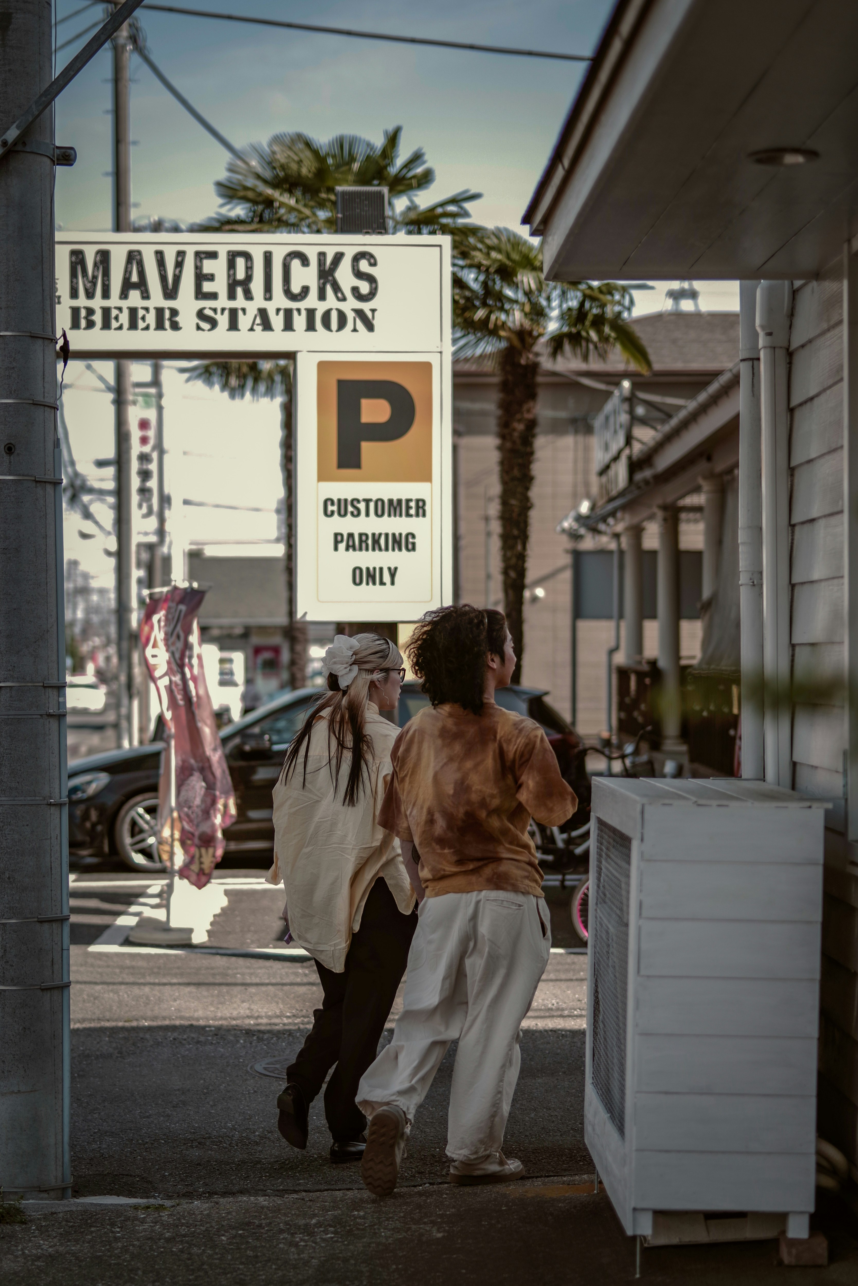 Two people walking by Mavericks Beer Station sign on a sunny street