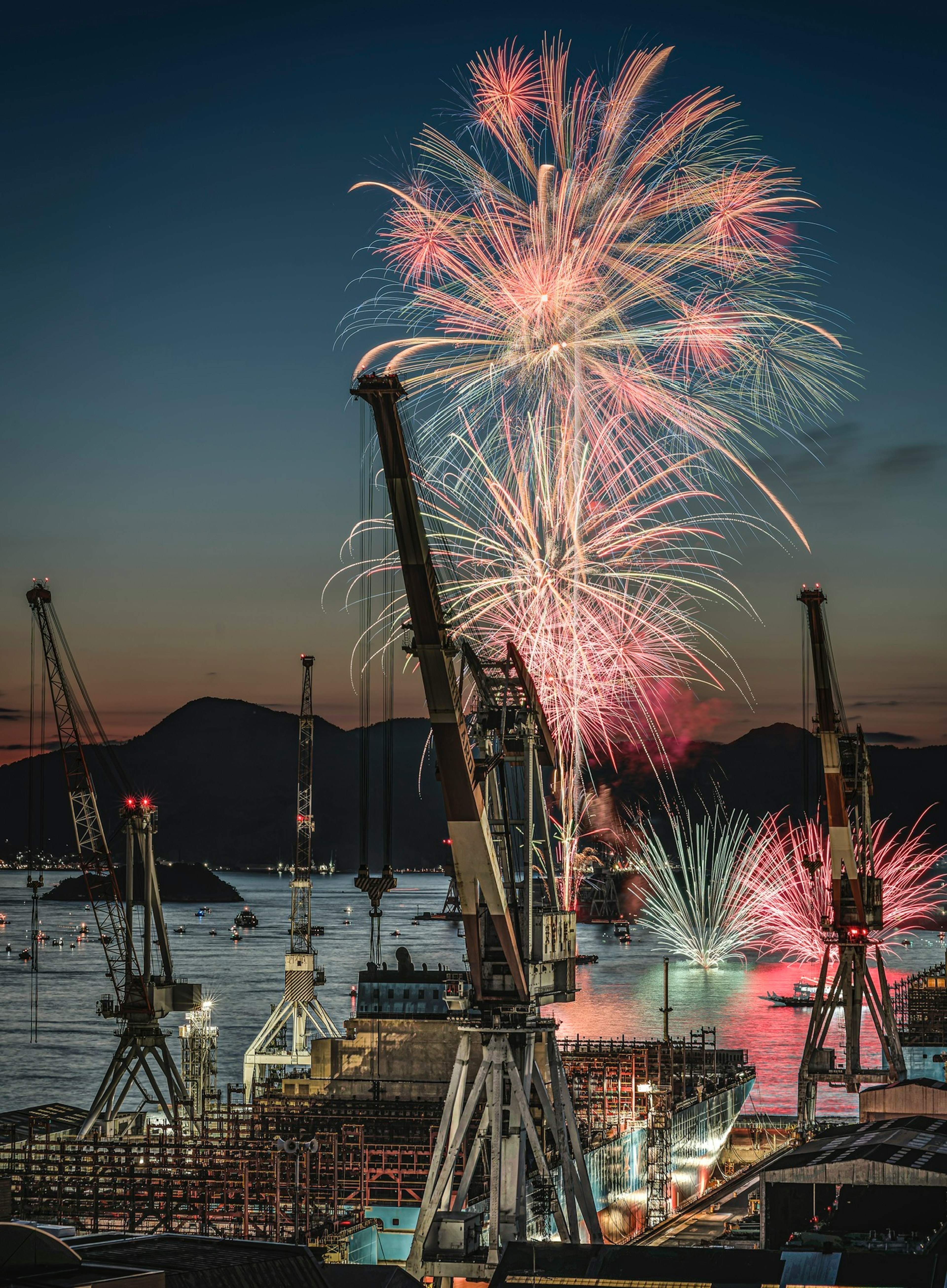 Fireworks lighting up the night sky over a harbor with crane silhouettes