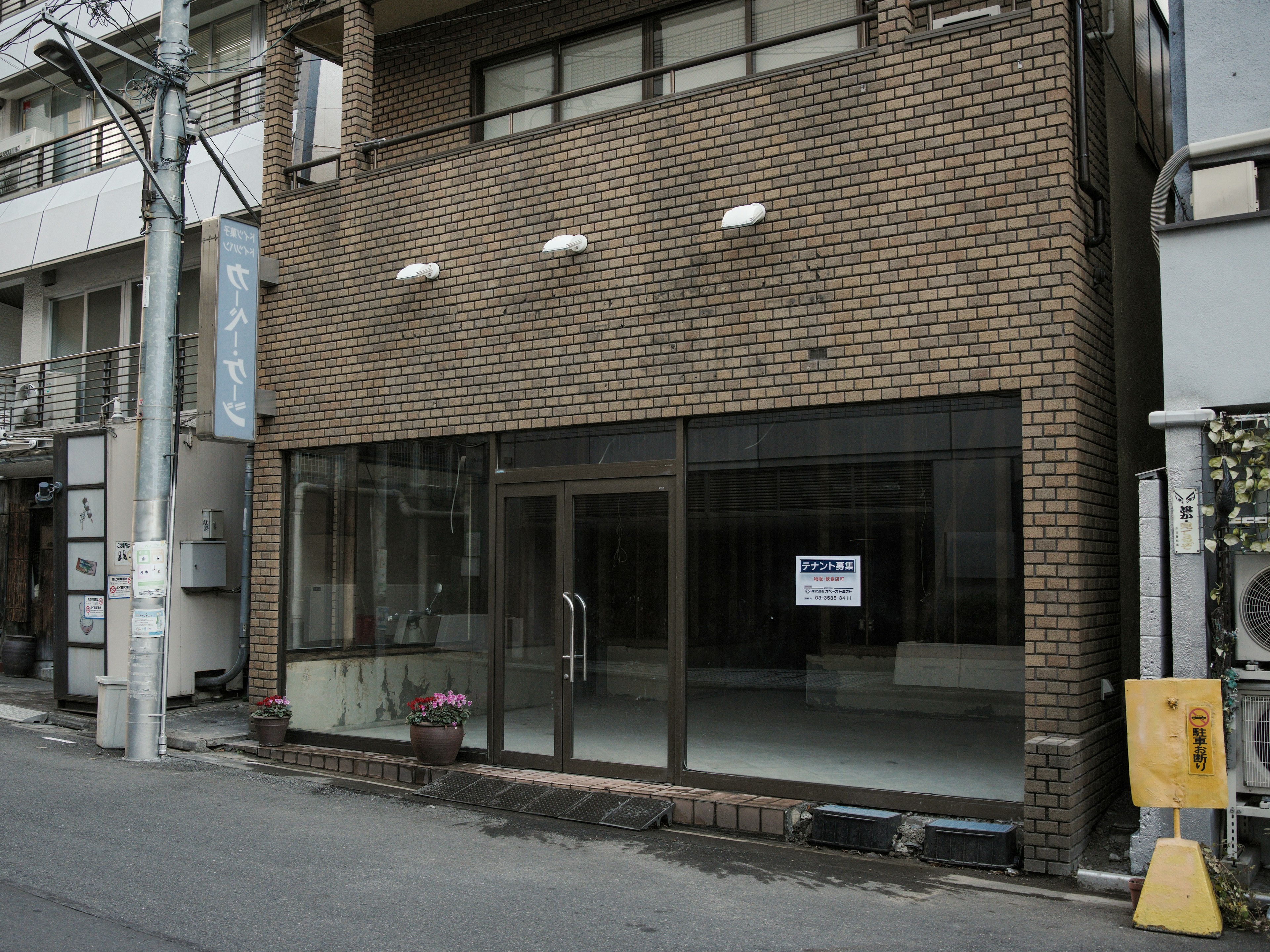 Exterior of a brick building with large windows A sign is visible inside the empty space Flower pot placed outside