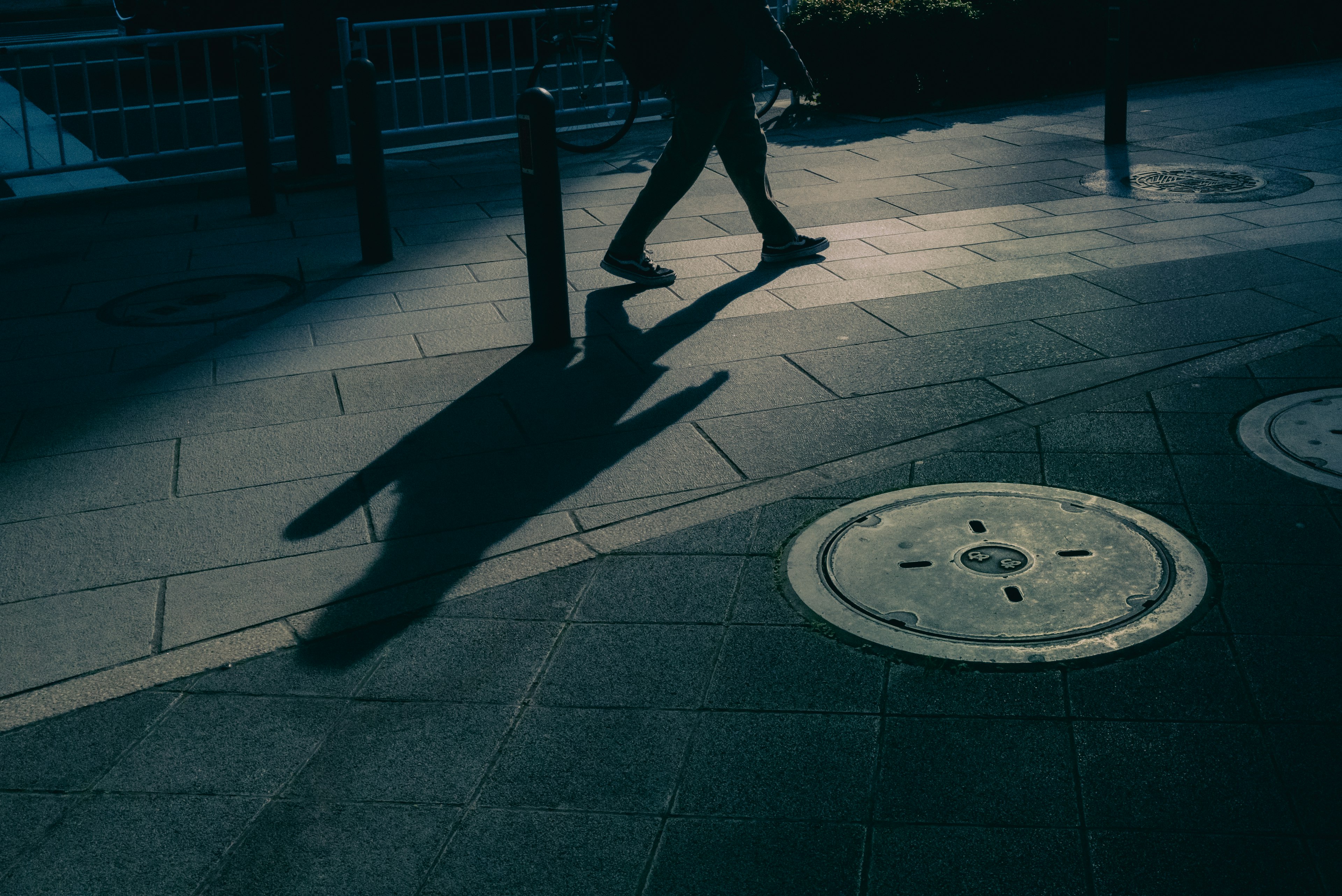 Una persona caminando con una sombra y una tapa de alcantarilla en un camino pavimentado