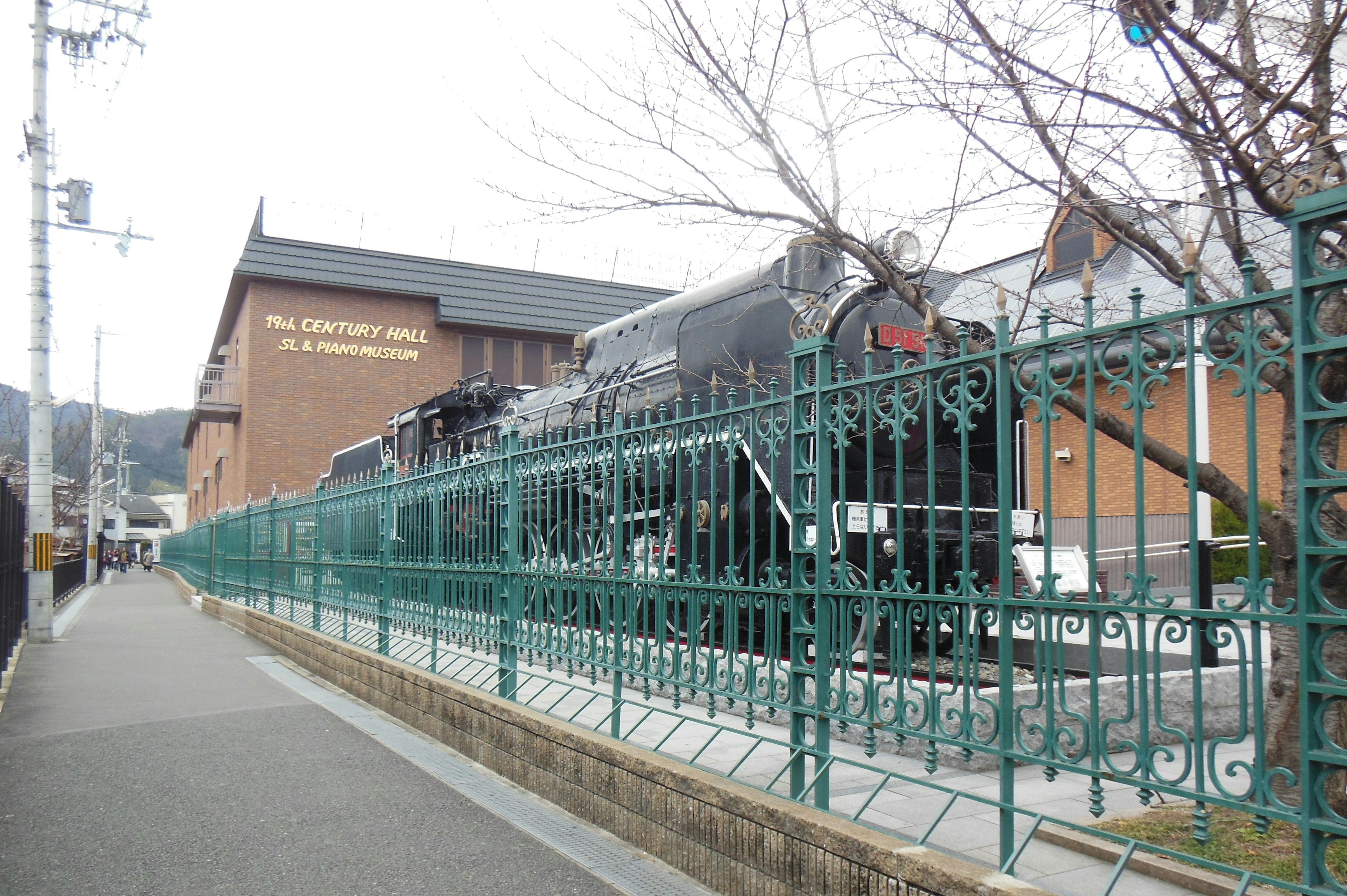 Steam locomotive beside a green fence with a railway museum in the background